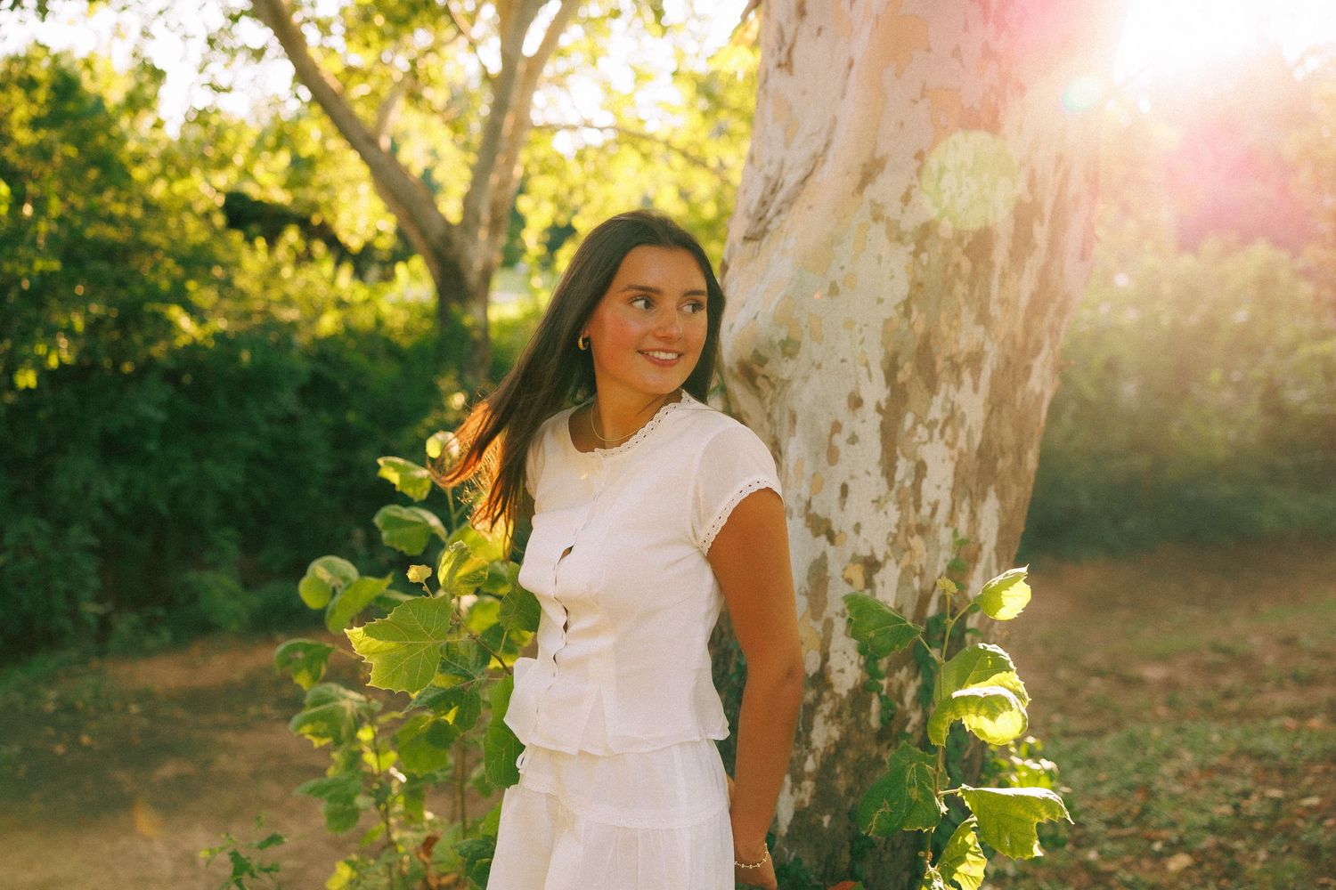 Young woman in white dress smiling beside a tree in a sunlit garden.