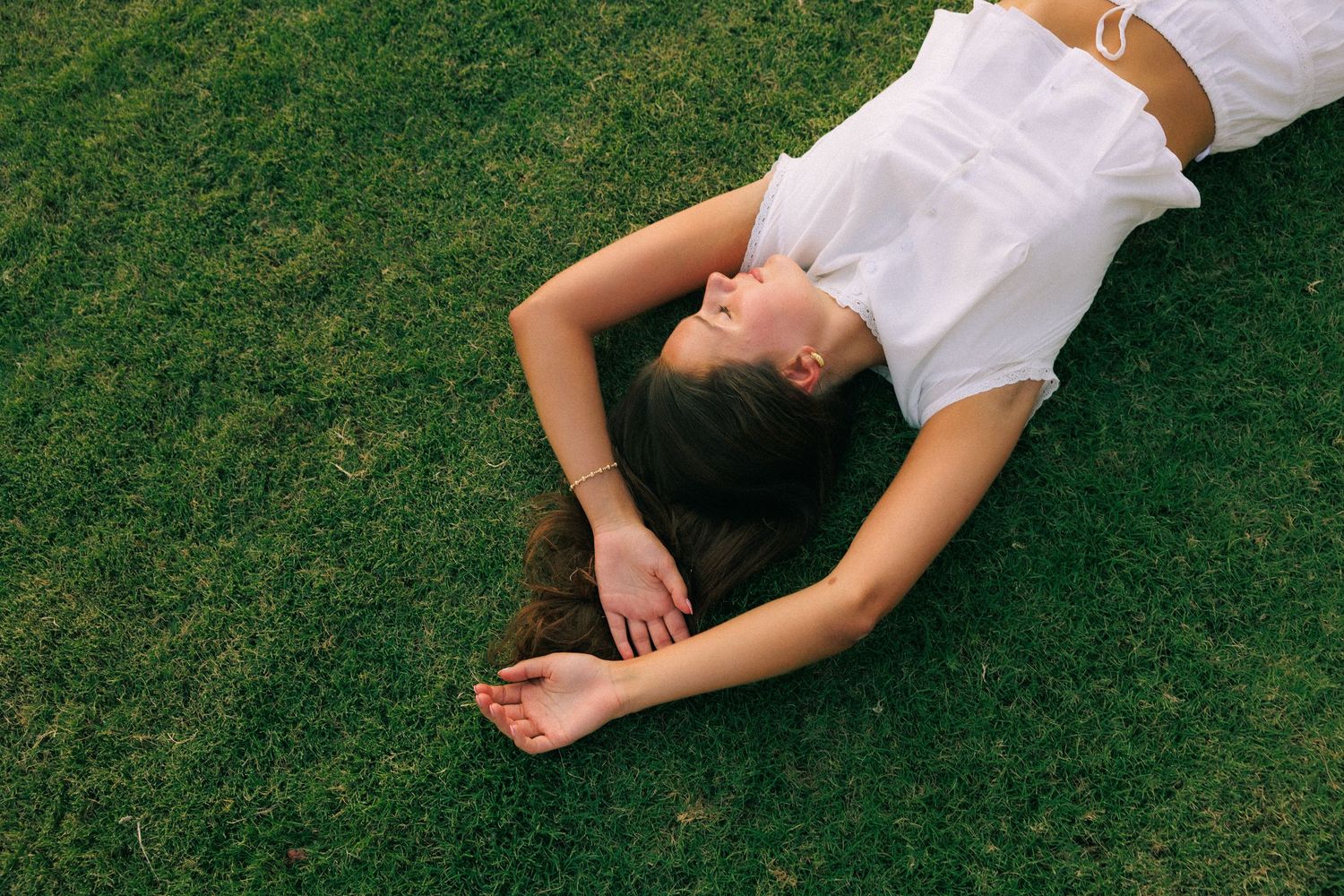 Young woman in white dress lying relaxed on lush green grass with eyes closed on a sunny day.