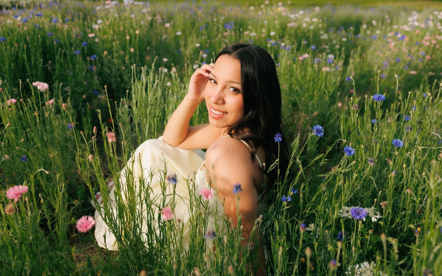 Smiling young woman in white dress sitting among wildflowers in a sunny meadow with pink and blue blooms.