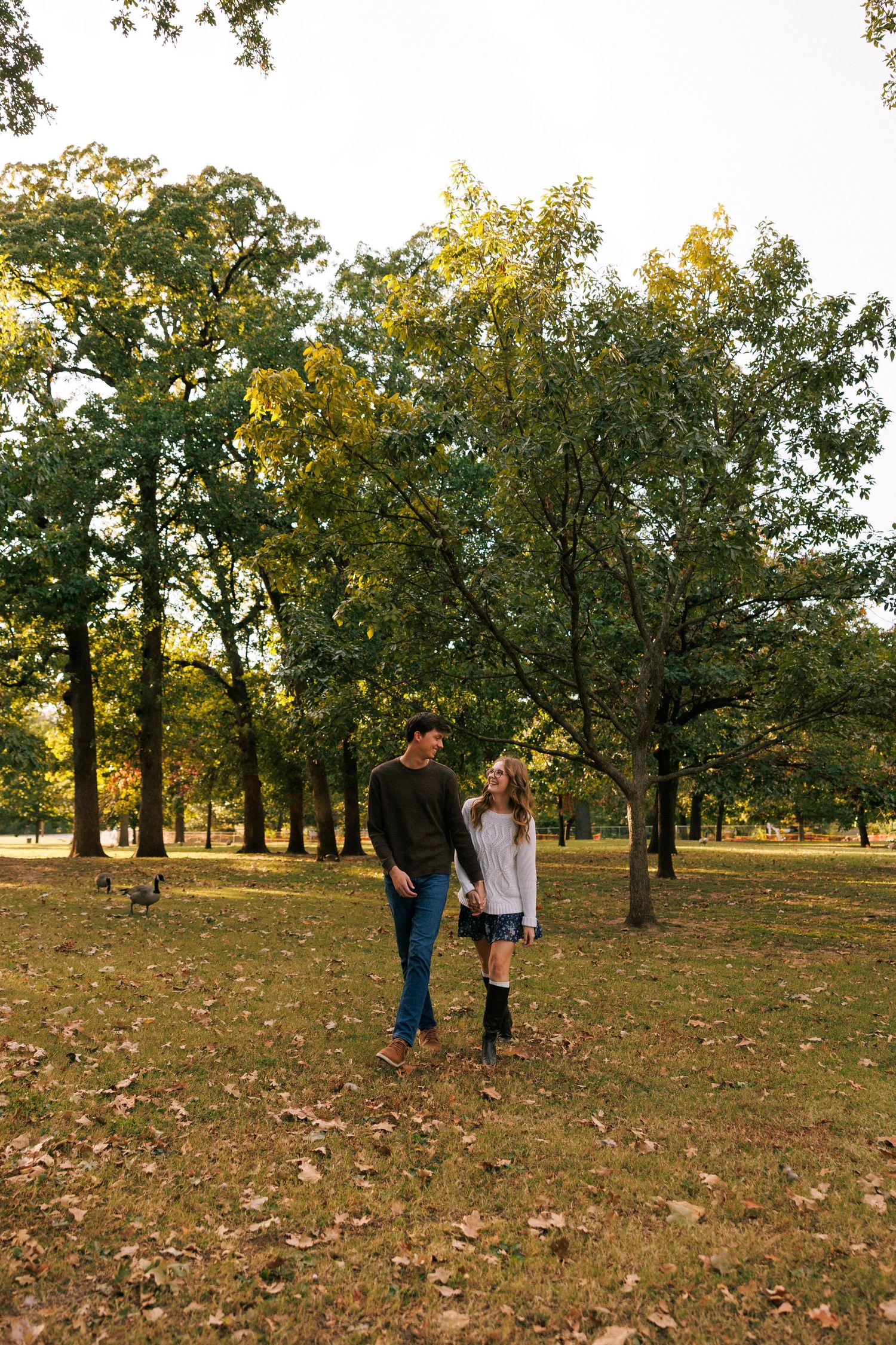 People walking through a park with large trees and scattered leaves on the ground.