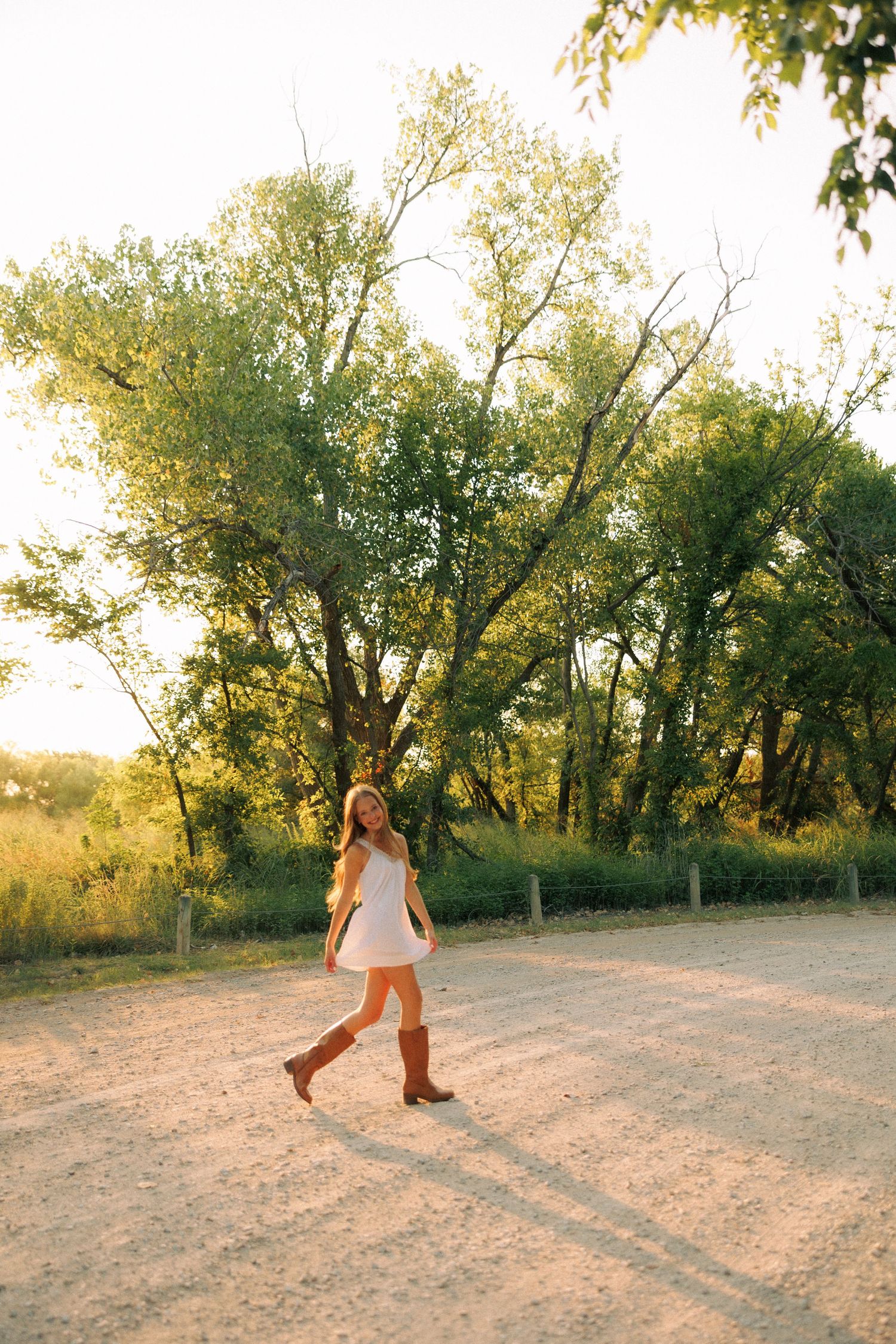 Young woman in white dress and cowboy boots walking on a sunny dirt road surrounded by lush green trees.