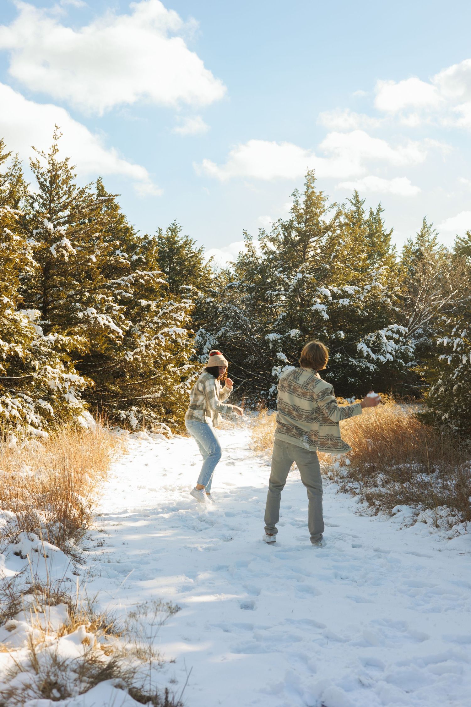 Two people playing and jumping in the snow surrounded by snow-covered evergreen trees.