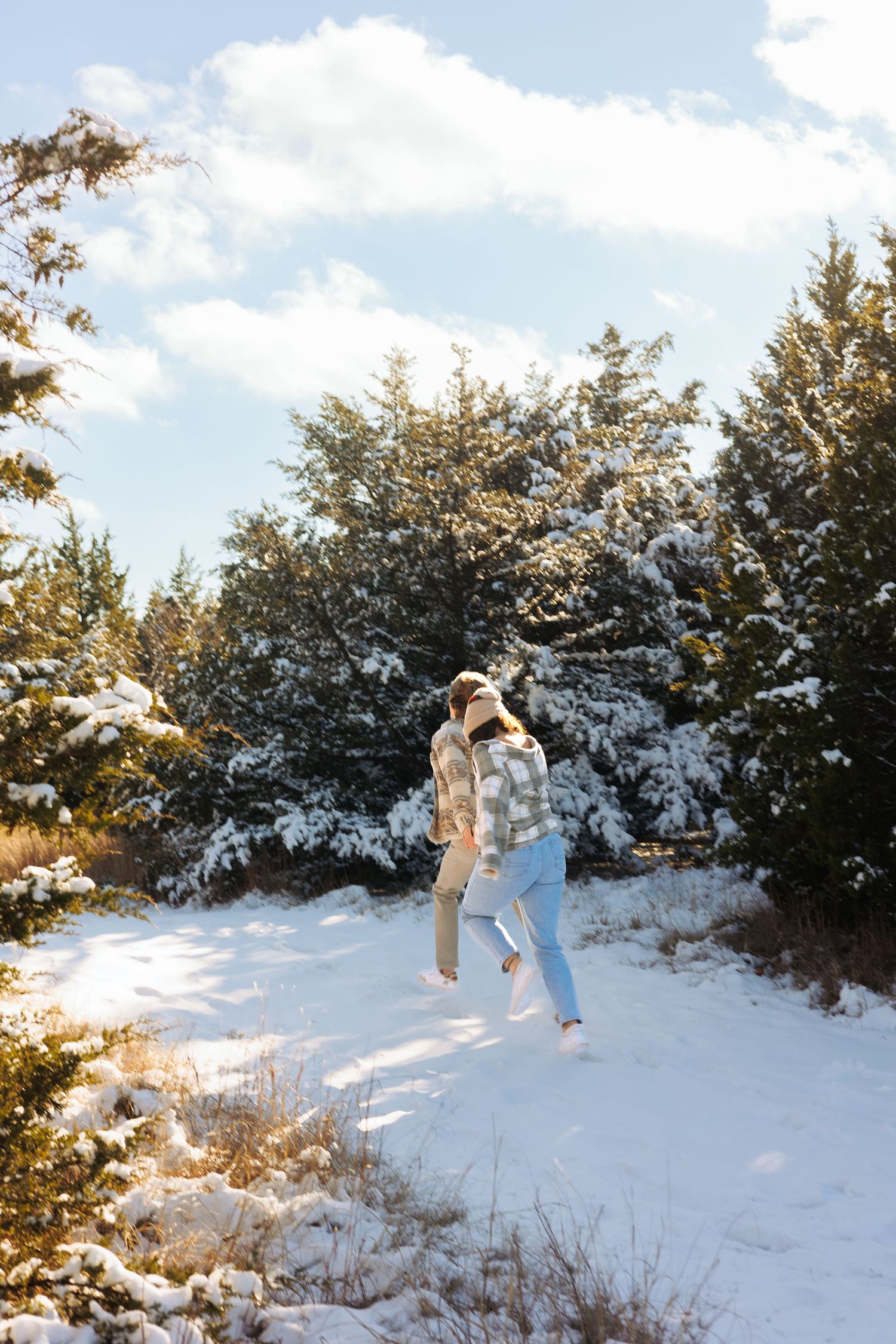 Two people playing in deep snow among snow-covered evergreen trees on a sunny winter day.
