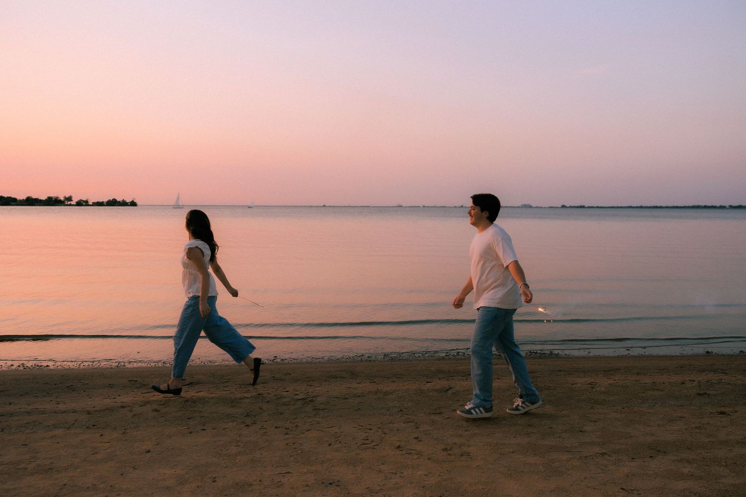 Couple walking along a sandy beach at sunset with pink and purple sky reflecting on calm water.