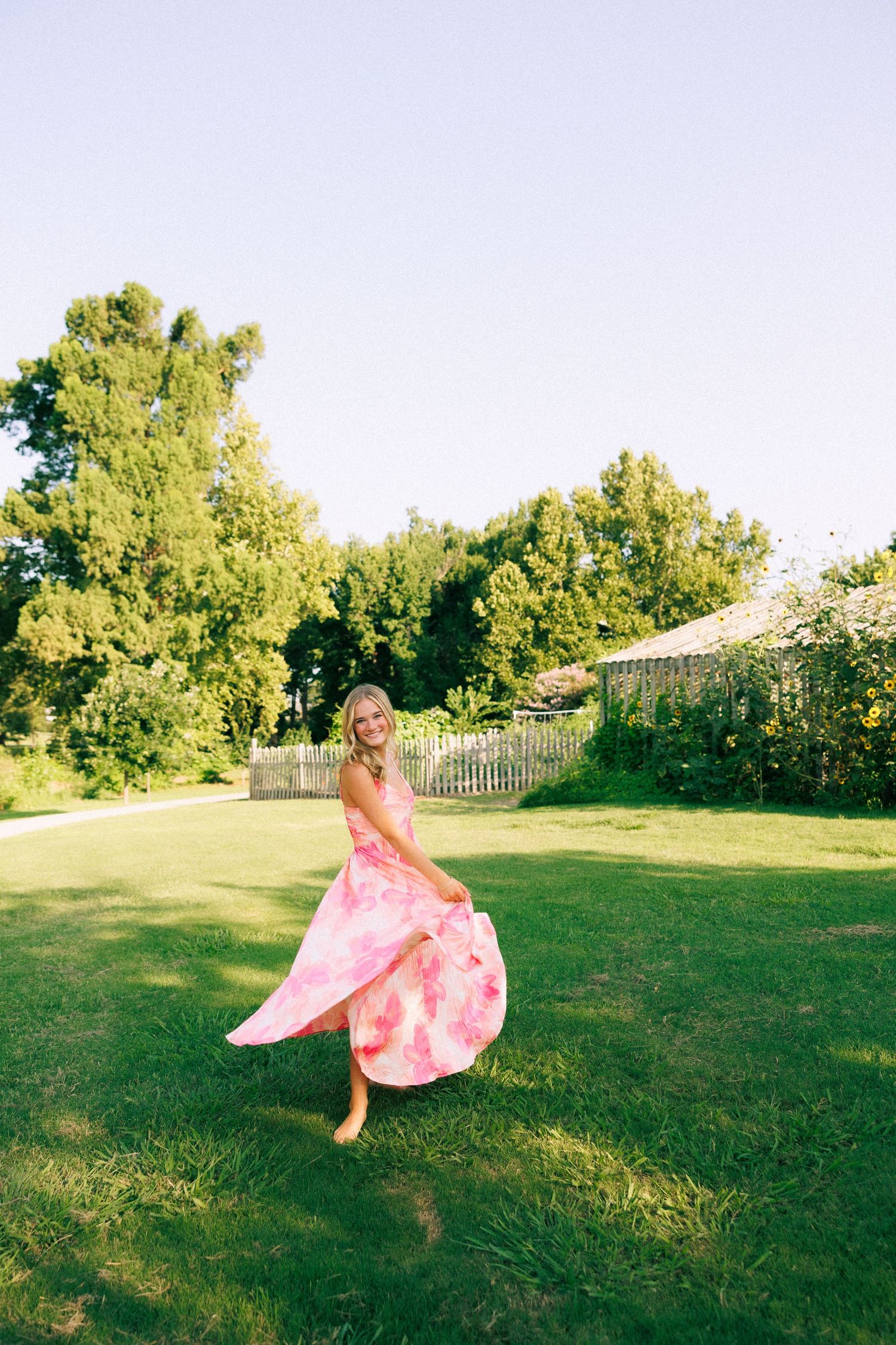 Woman in flowing pink floral maxi dress twirling barefoot on lush green lawn with trees and white fence backdrop.