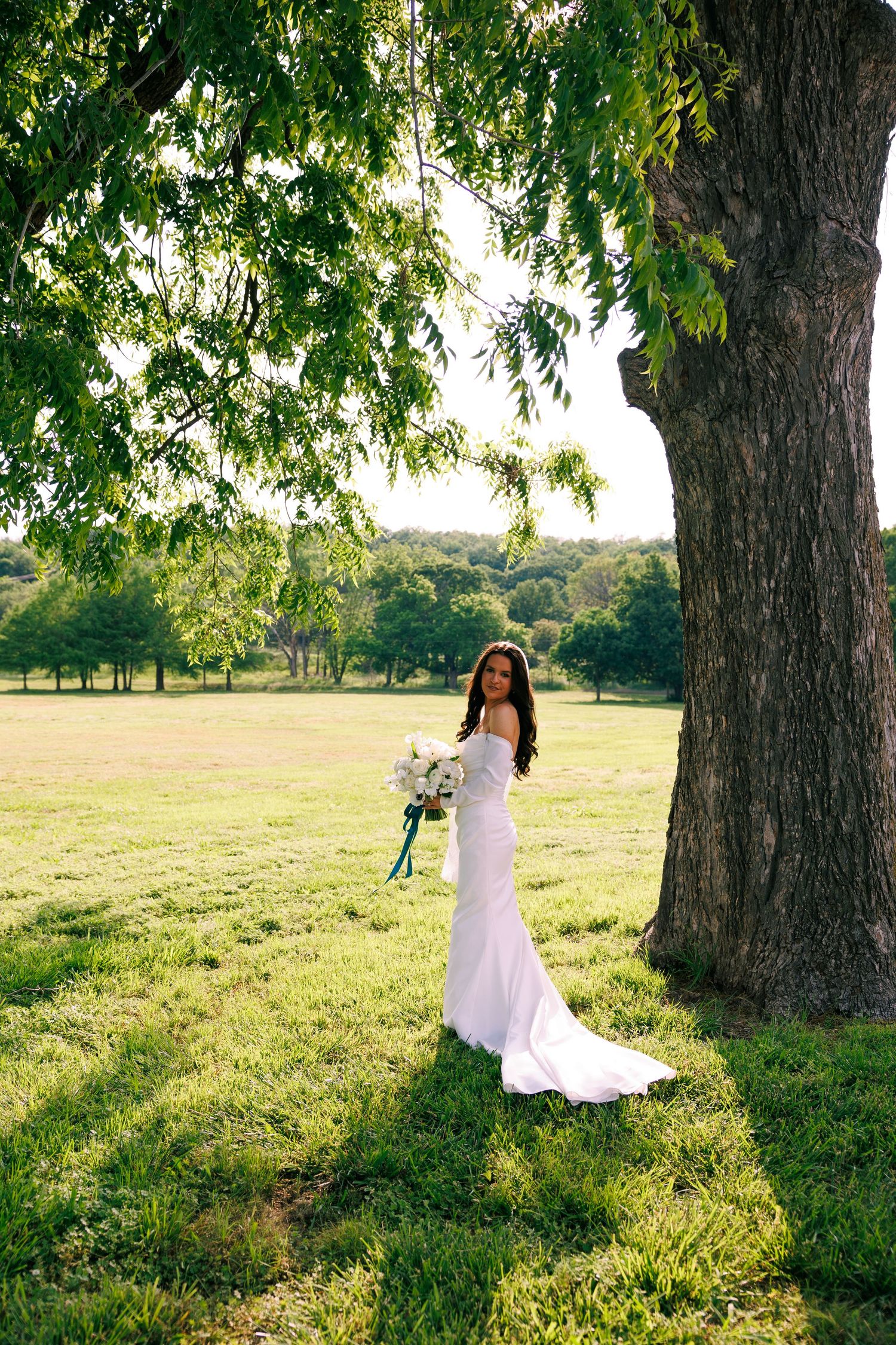 Bride in white dress poses gracefully under large trees in a sunlit meadow.