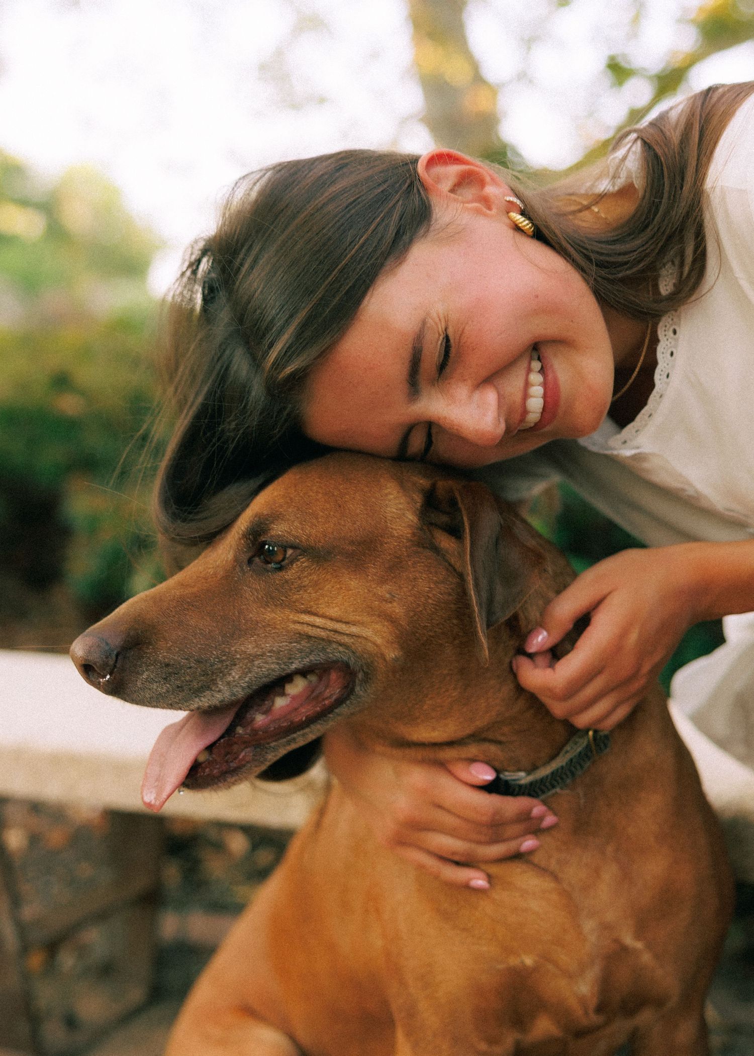 A smiling young woman with brown hair hugs a large brown Rhodesian Ridgeback dog outdoors.