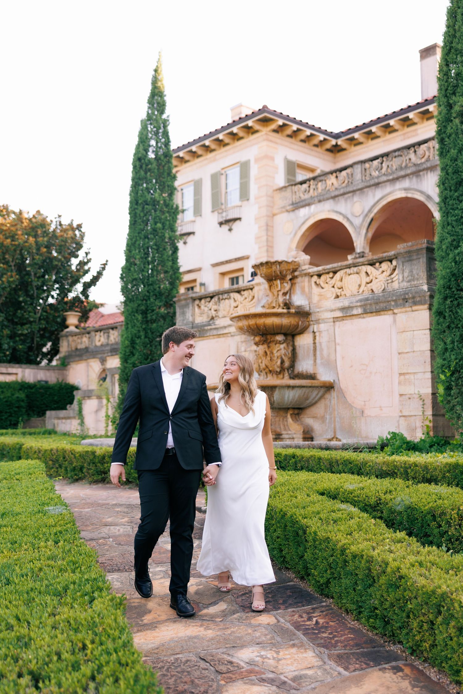 Couple walks through Mediterranean-style garden with stone fountain and cypress trees.