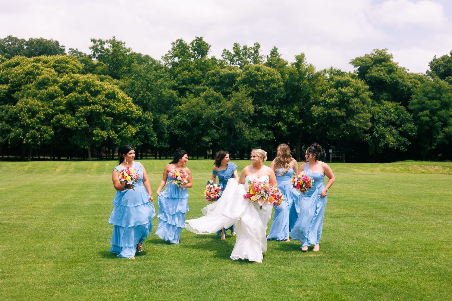 Wedding party in light blue dresses celebrates on a sprawling green lawn.