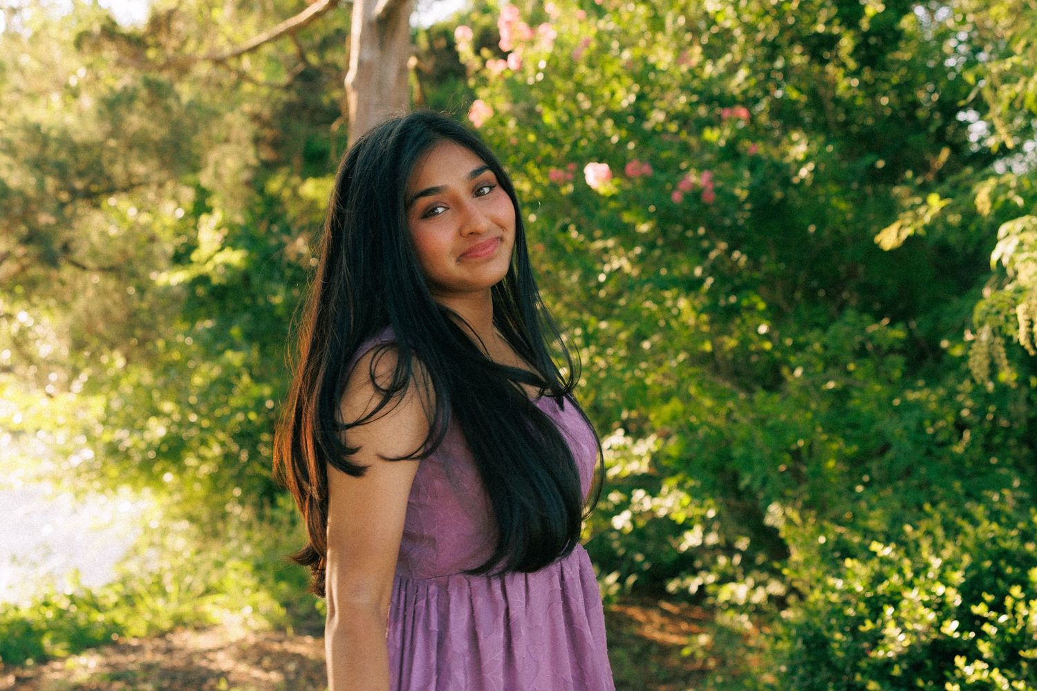 Young woman in purple dress leaning against tree in lush green garden, long dark hair, natural outdoor setting.