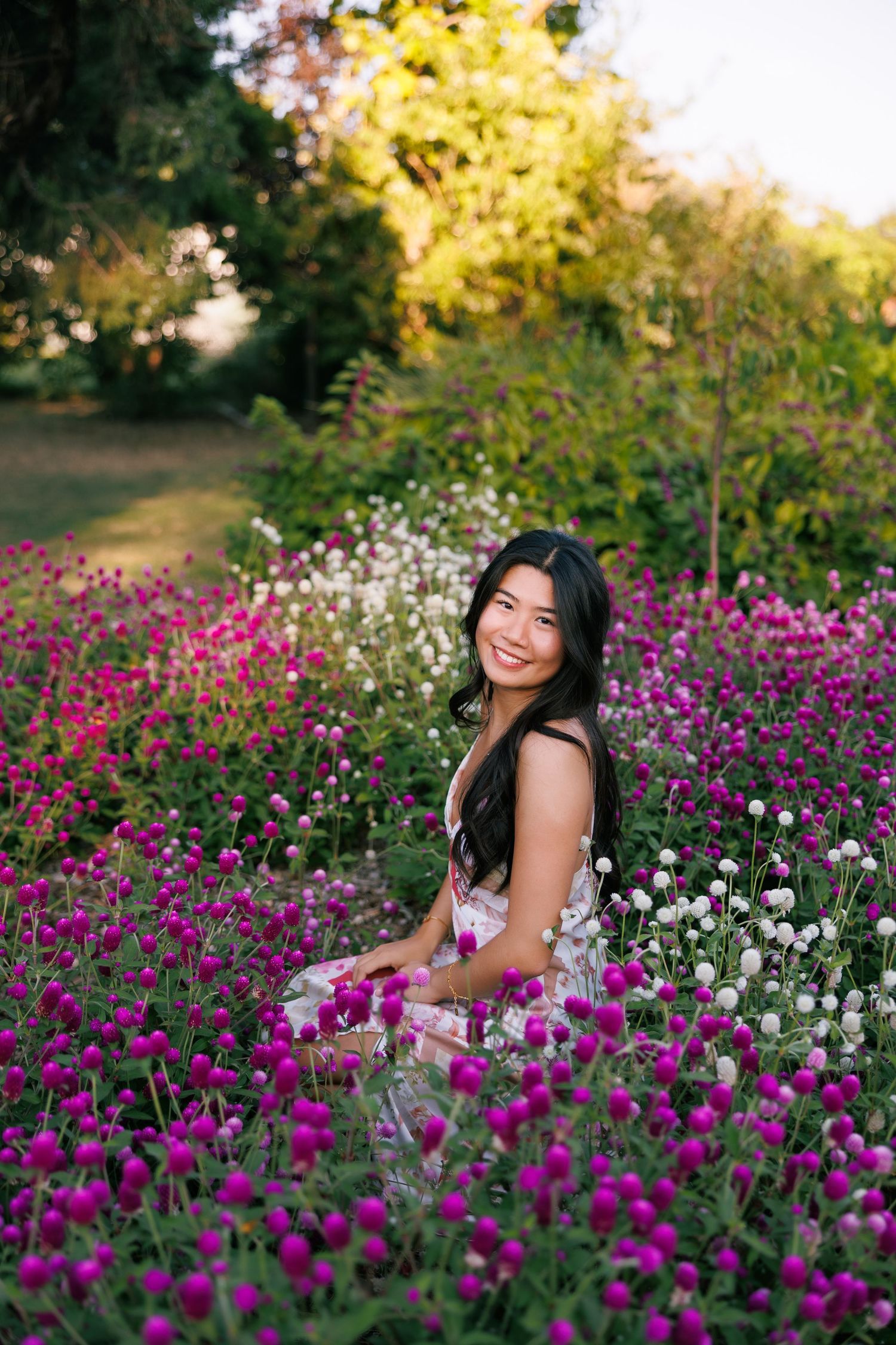 Person in dark dress poses among vibrant purple and white flowers in garden at sunset.