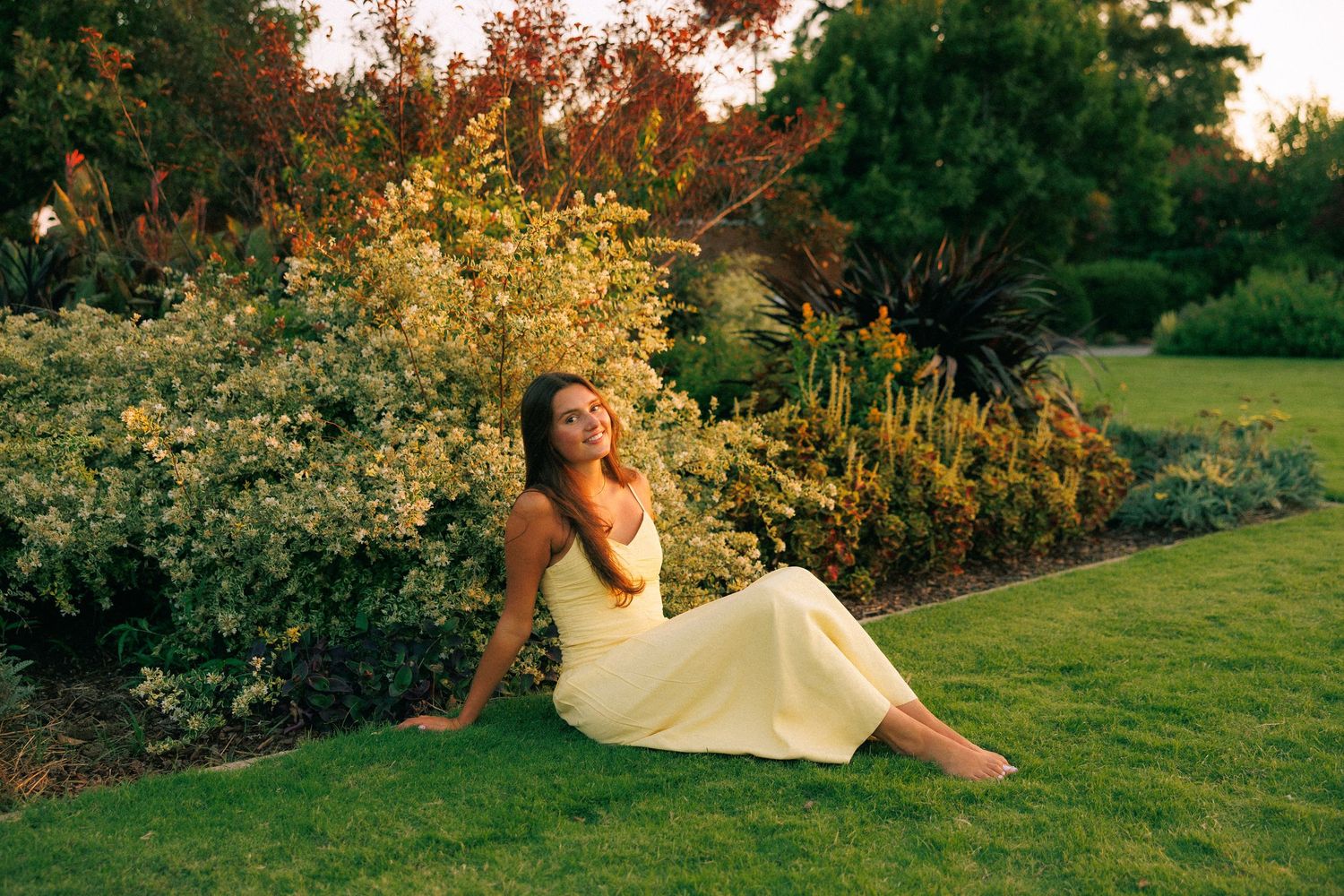 Woman in yellow dress sitting on lush green grass beside colorful garden shrubs in warm golden sunset light.