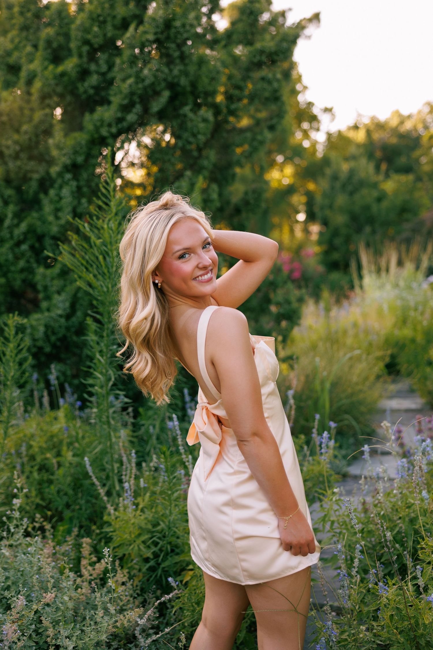 Woman in white dress poses playfully in a garden setting during golden hour.