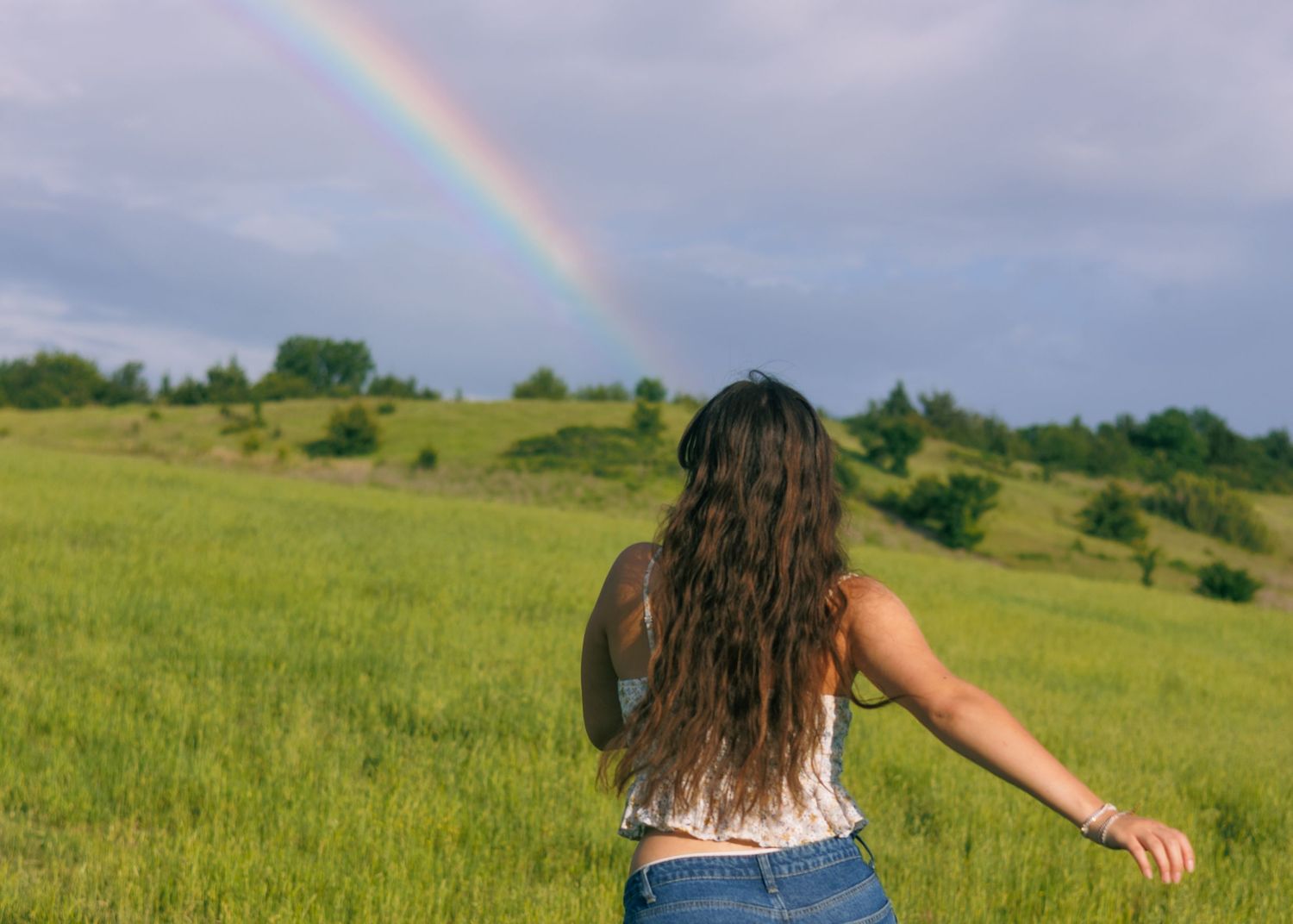 A person with long hair stands in a meadow with arms outstretched beneath a rainbow.