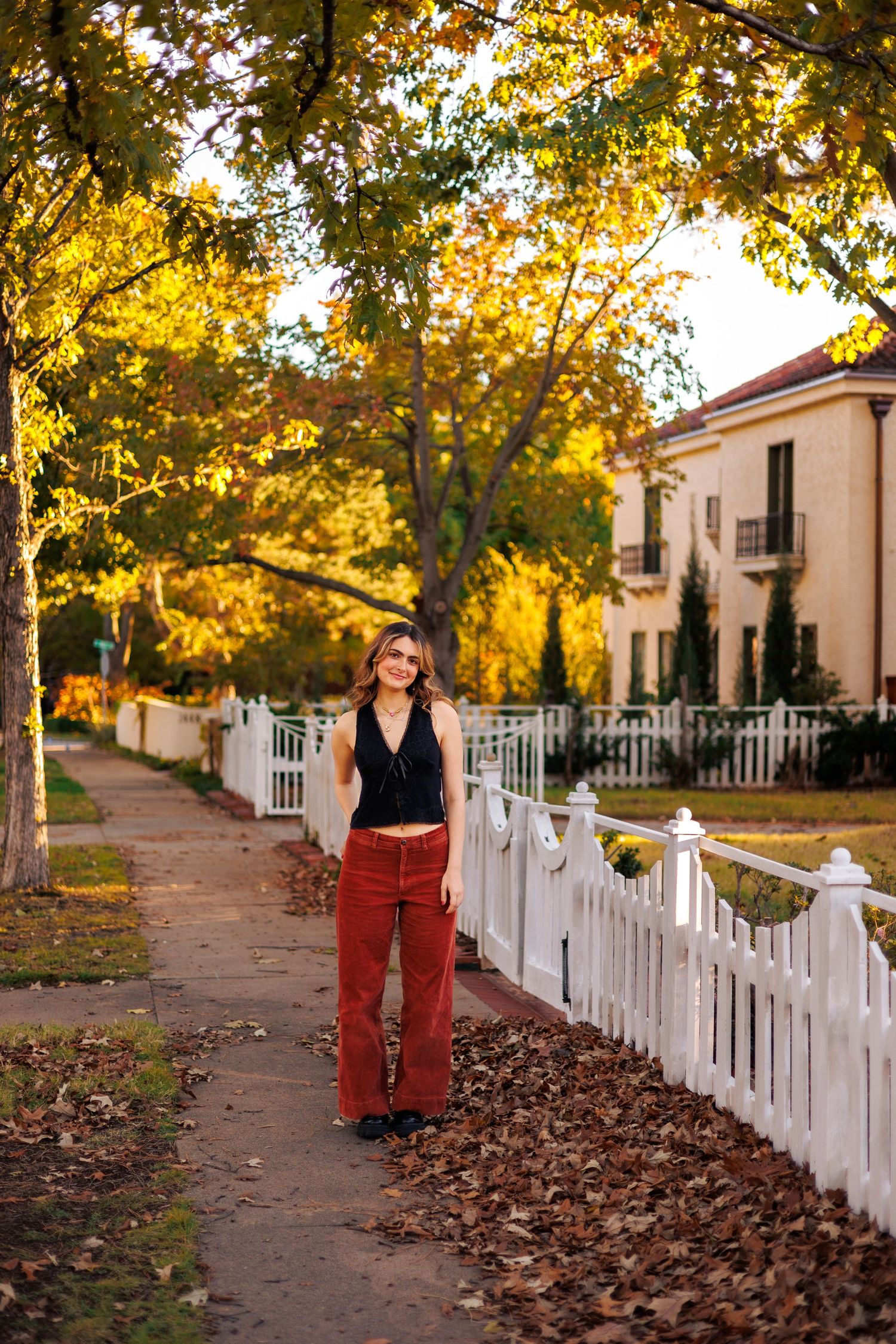 White picket fence lines a leaf-covered walkway with autumn trees and buildings.