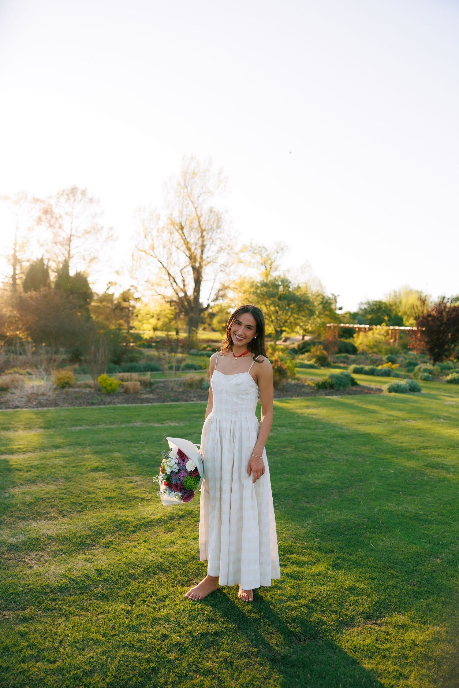 Woman in white sundress holding colorful bouquet standing barefoot on lush green lawn at golden hour.