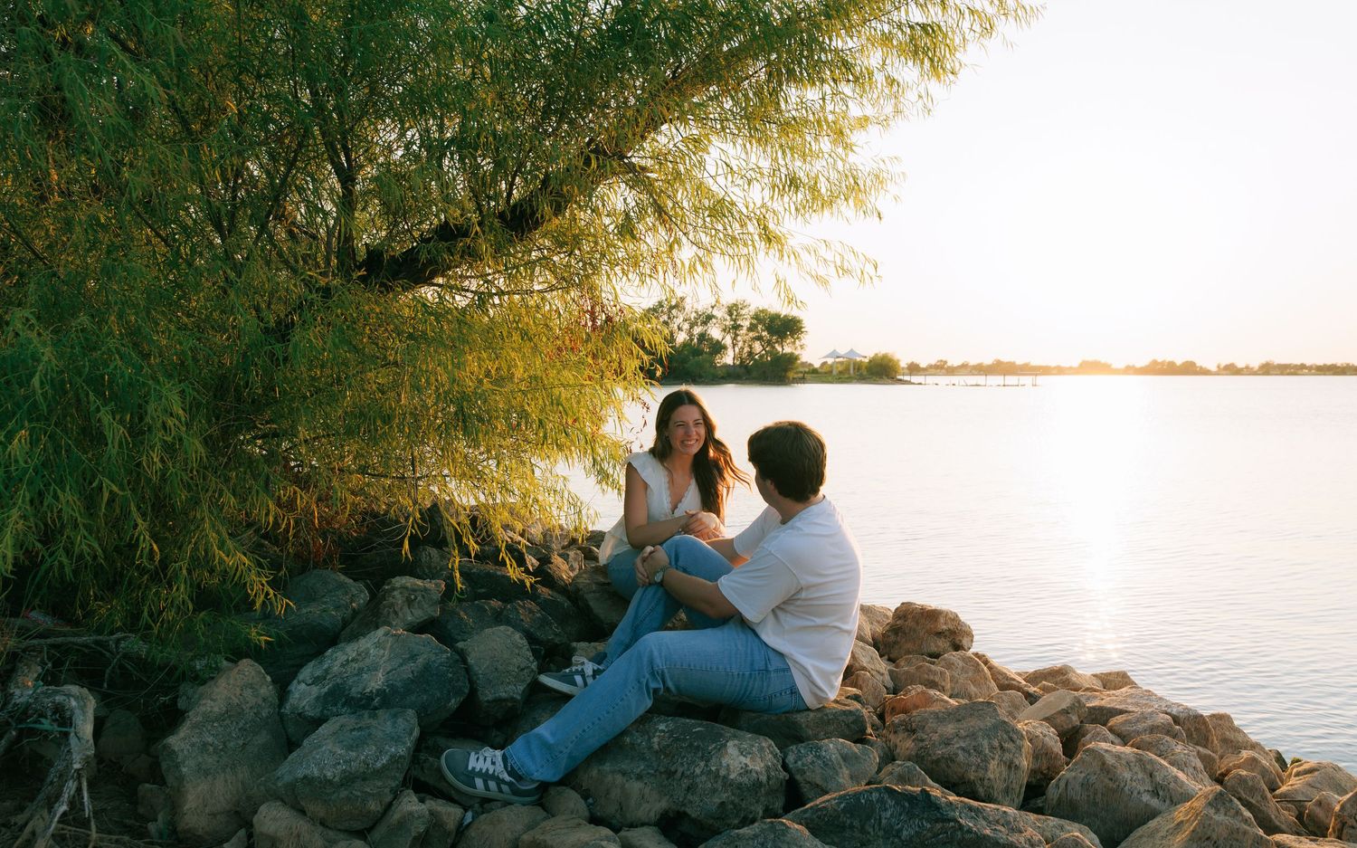 Couple sitting on rocks by a serene lake at sunset, surrounded by lush green trees in a romantic outdoor setting.