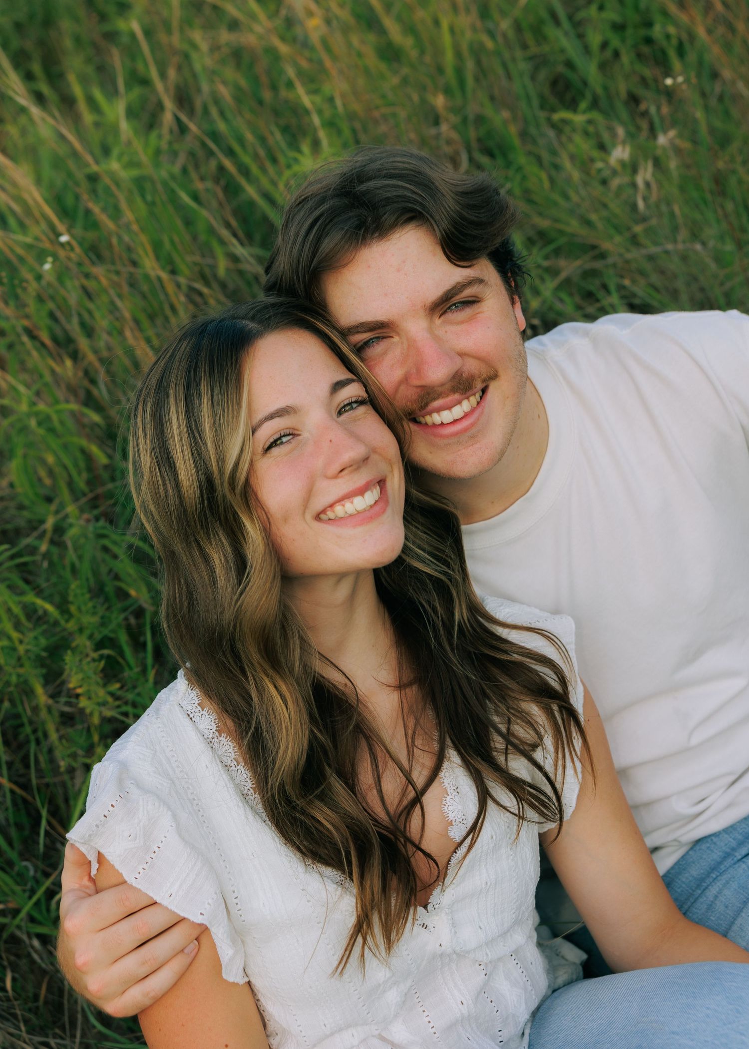 Happy young couple smiling and embracing outdoors in a green field, wearing casual white tops.