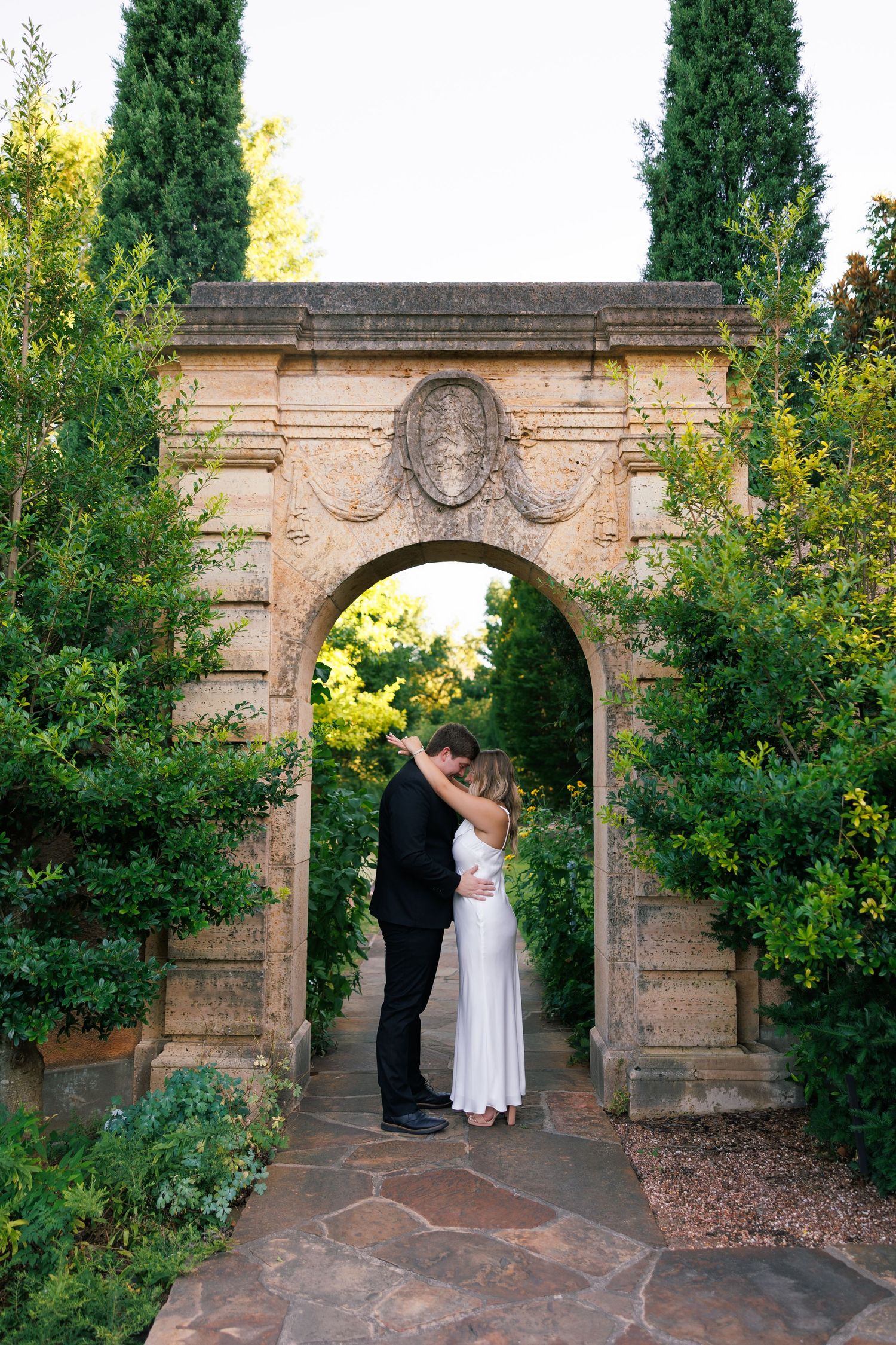 A couple shares an intimate moment under an ornate stone garden archway.