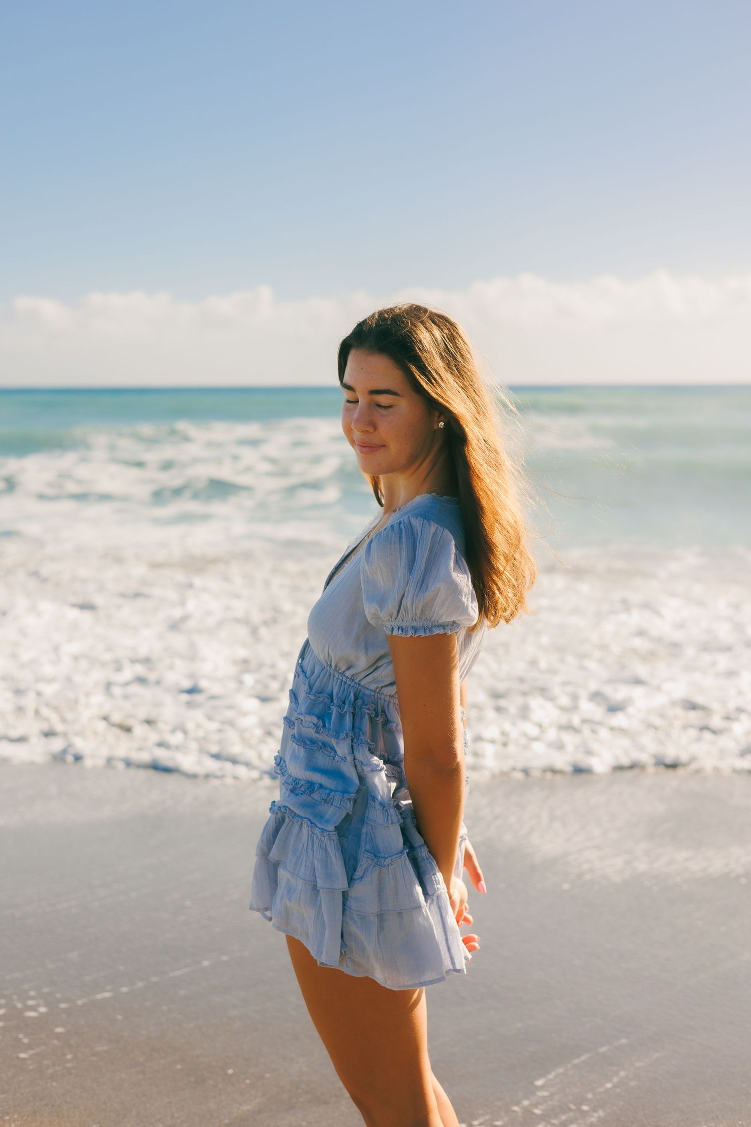 A light blue ruffled dress flows in the ocean breeze as waves crash on a sandy beach.