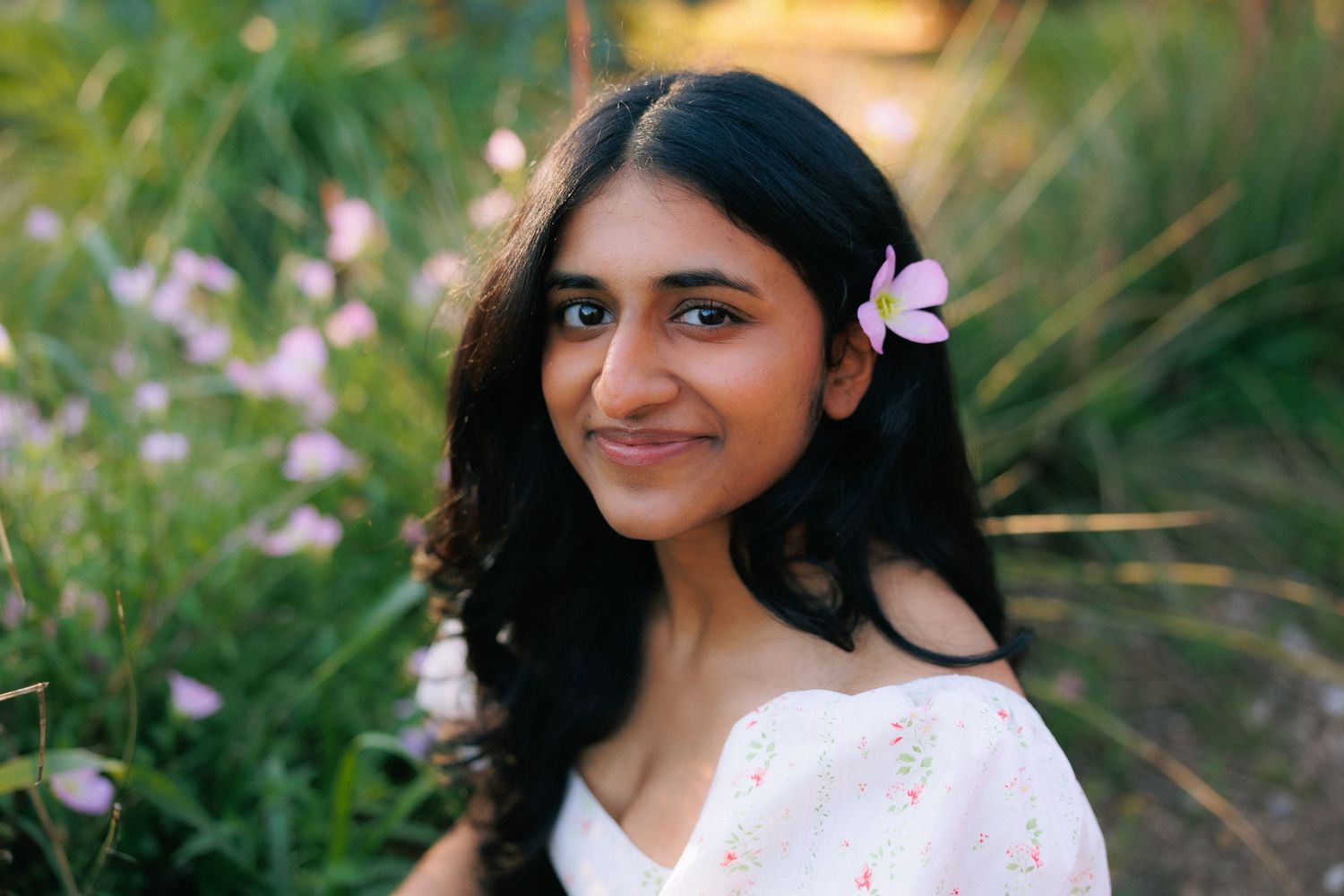 Young woman with a pink flower in her hair smiling outdoors among green plants and pink flowers.