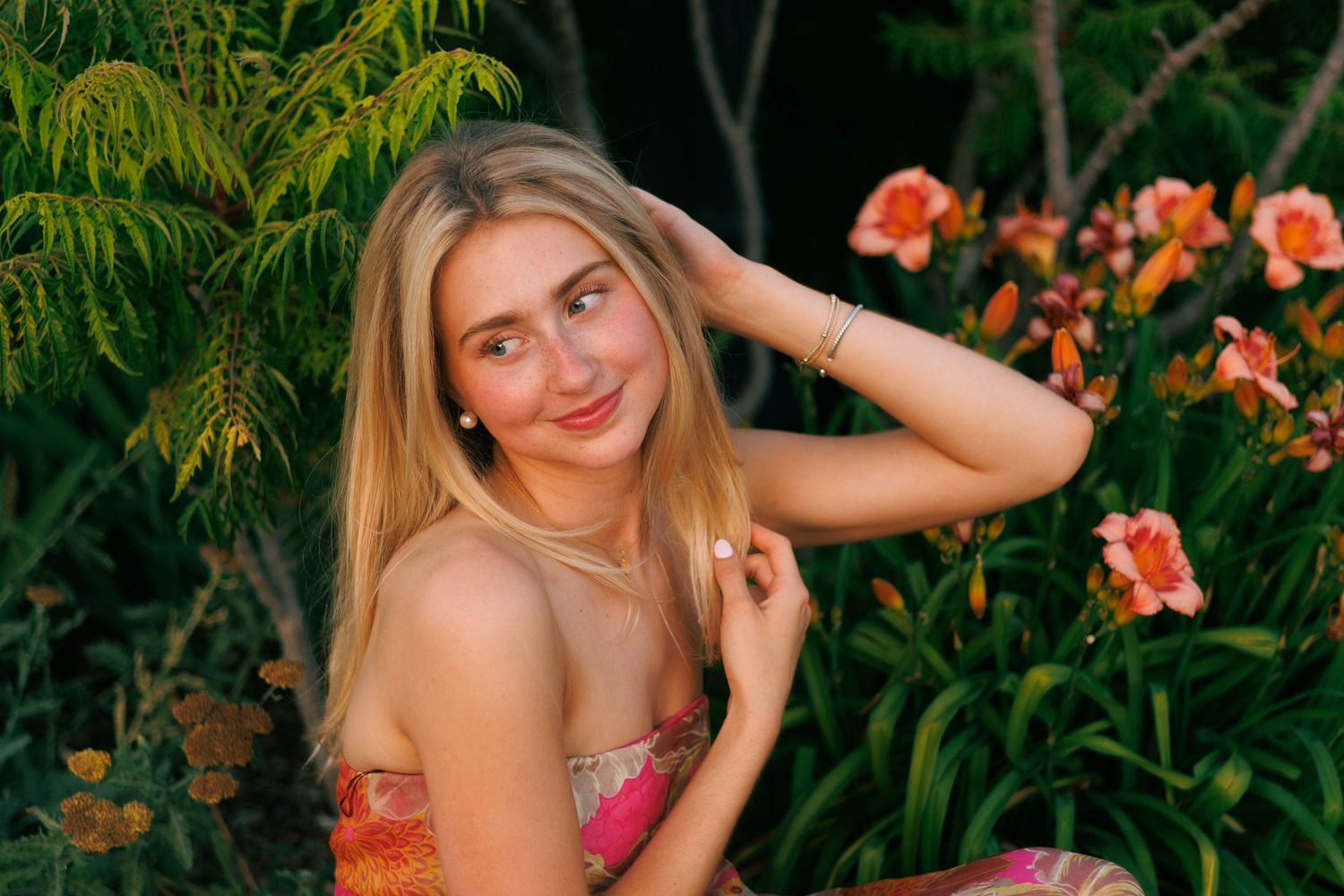 Young blonde woman in floral top posing among orange flowers and green foliage in a garden setting.