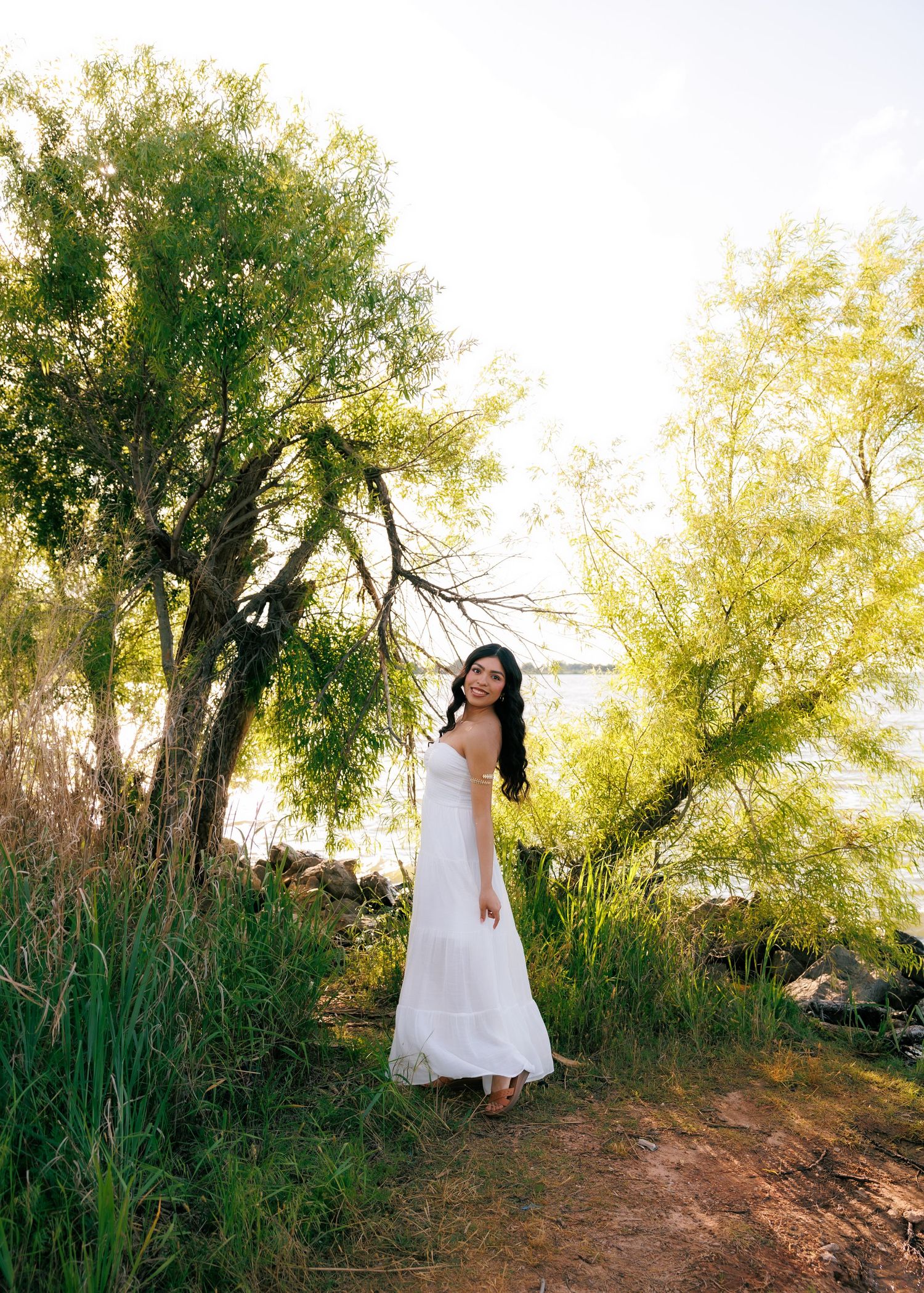 Person in white dress poses on dirt path surrounded by willow trees and tall grasses.