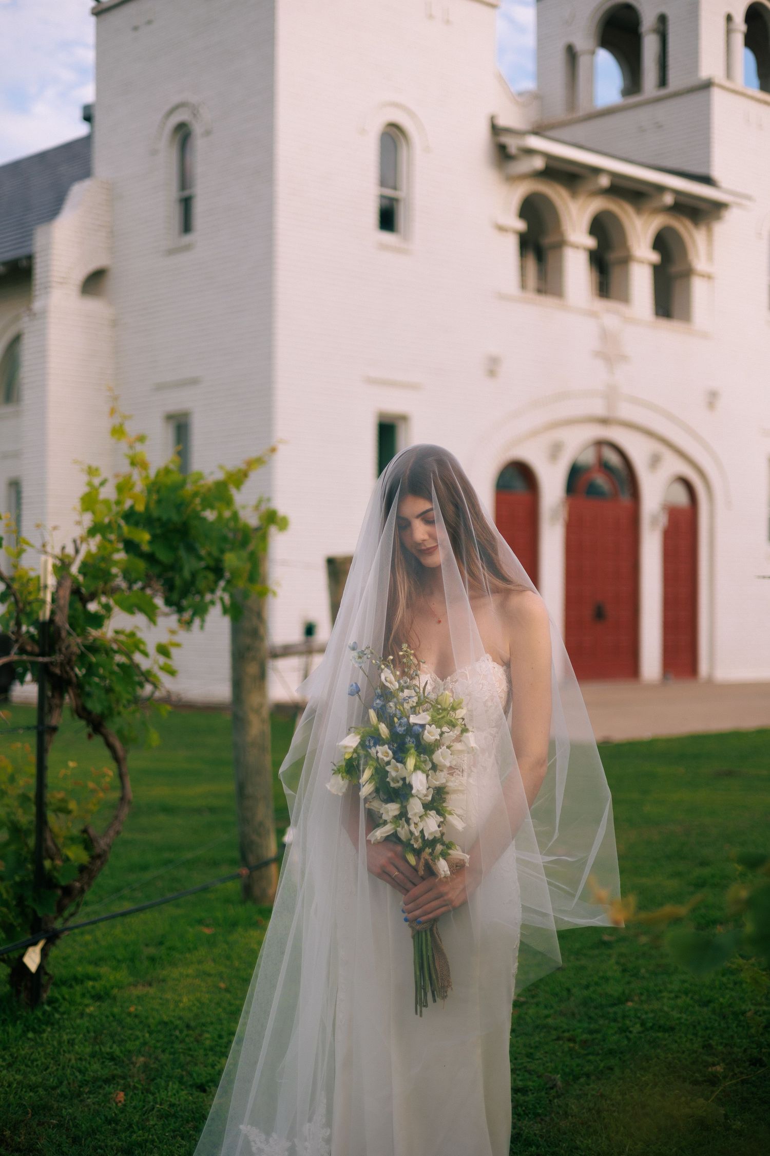 A bride in a flowing veil poses in front of a white mission-style church building.