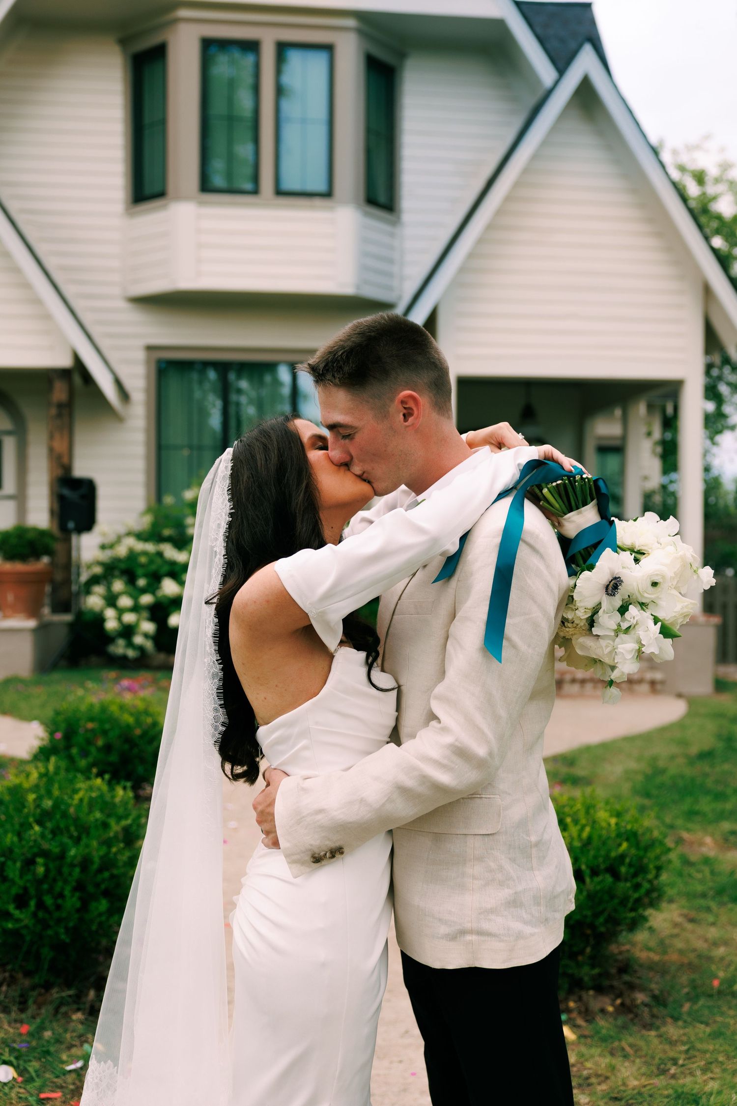 Newlyweds embrace in front of a white house during their outdoor wedding photoshoot.