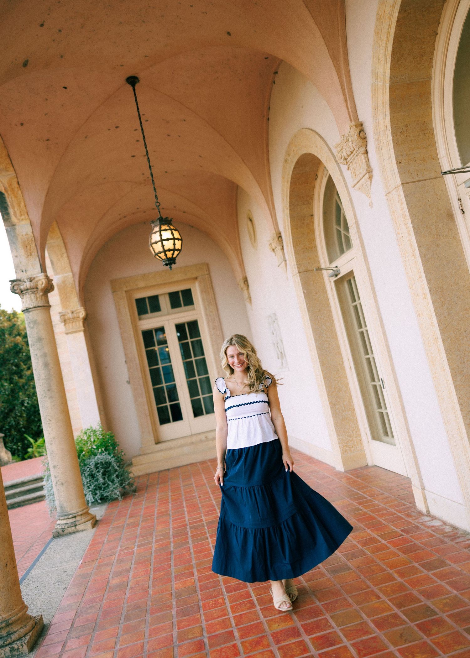 Woman in white top and navy maxi skirt posing on terracotta tile loggia with arched columns.