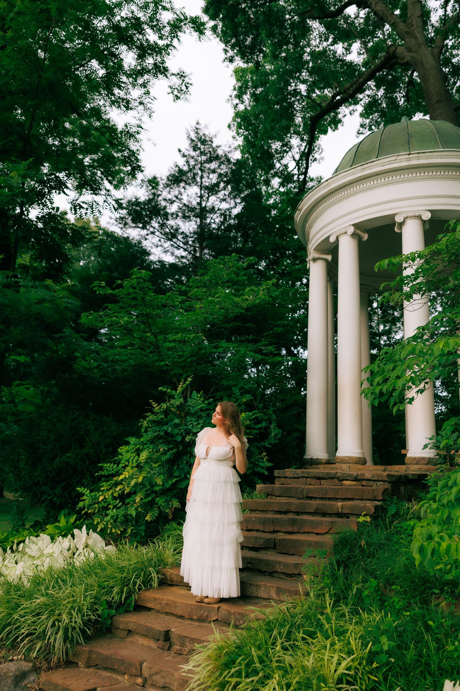 Bride in white tiered gown stands on stone steps beside a classic white rotunda surrounded by lush green gardens.
