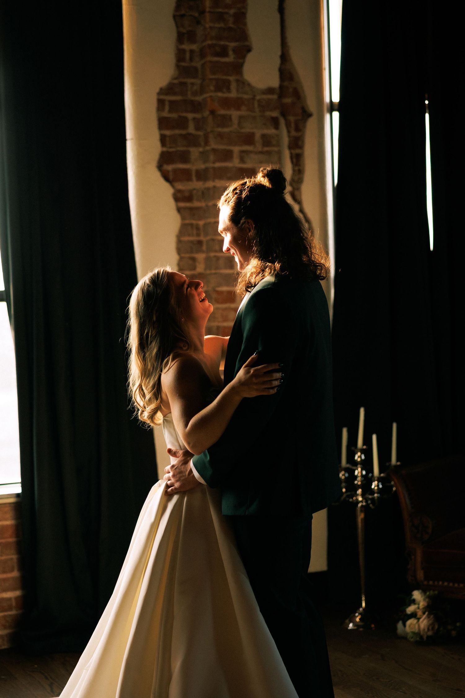 Silhouetted wedding couple dancing in dramatic window lighting against exposed brick walls.