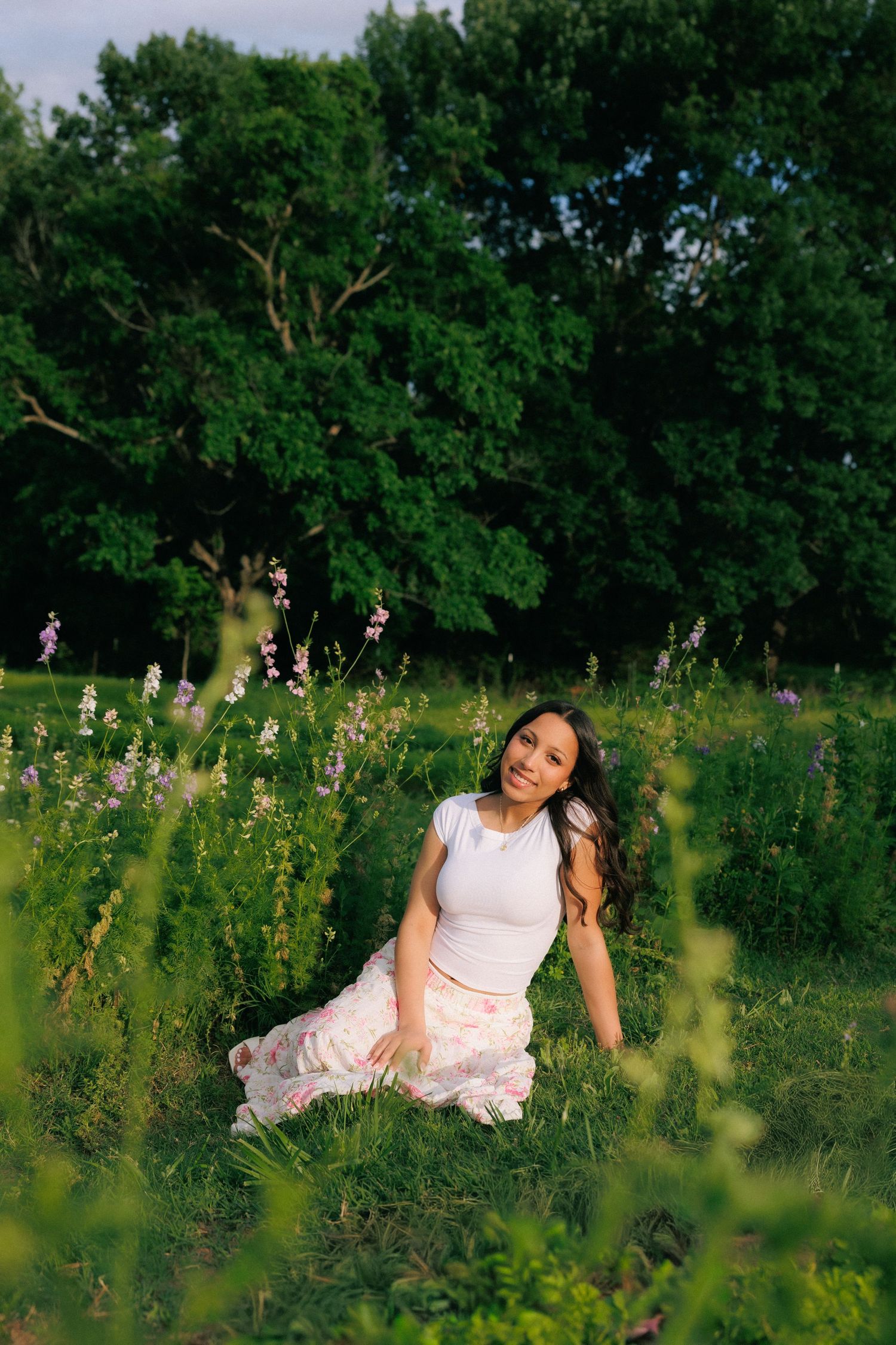 Young woman in white top and floral skirt sitting gracefully in a lush green meadow with wildflowers at golden hour.