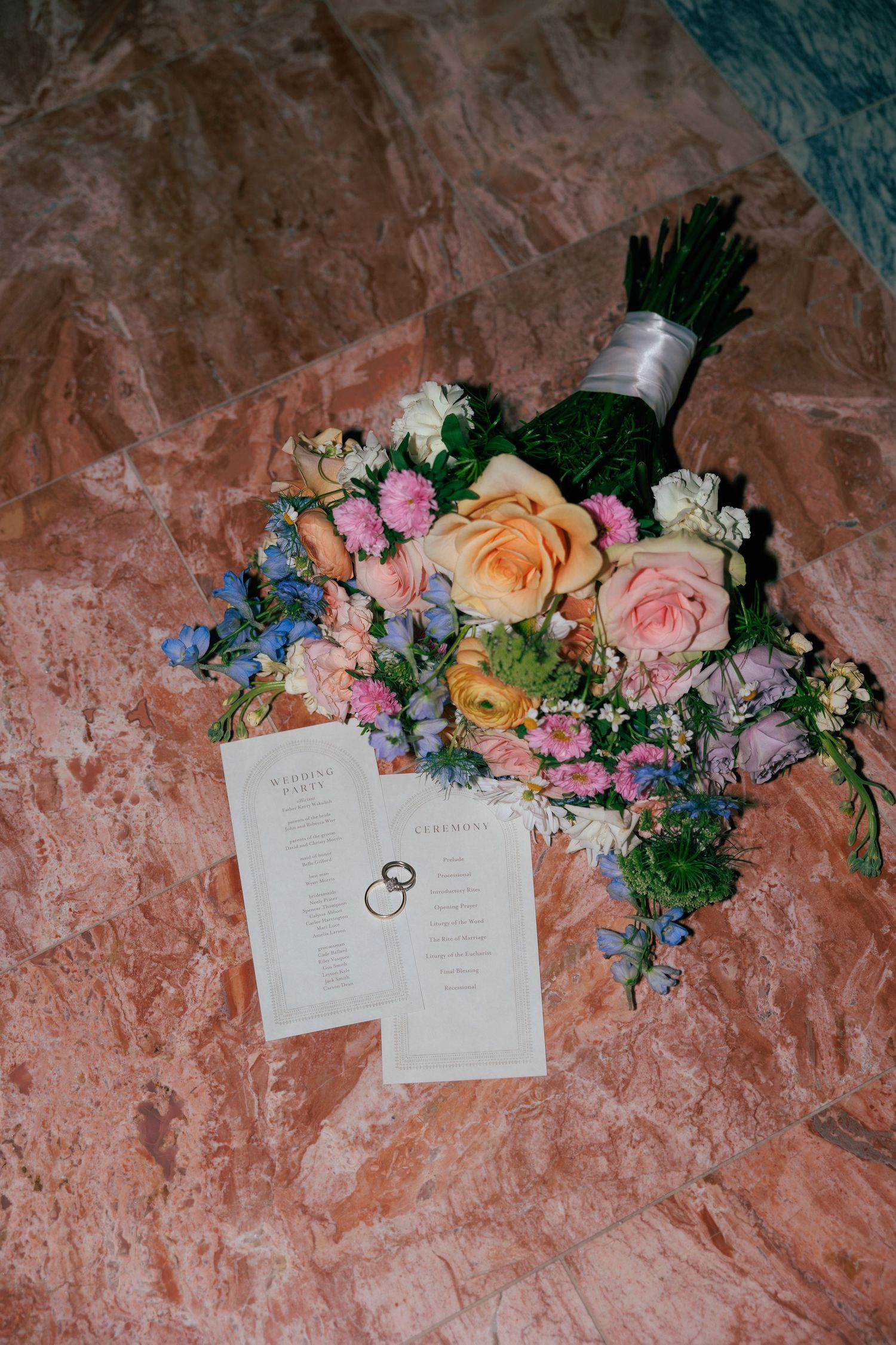 Wedding stationery and rings styled with pastel flowers against textured coral background.