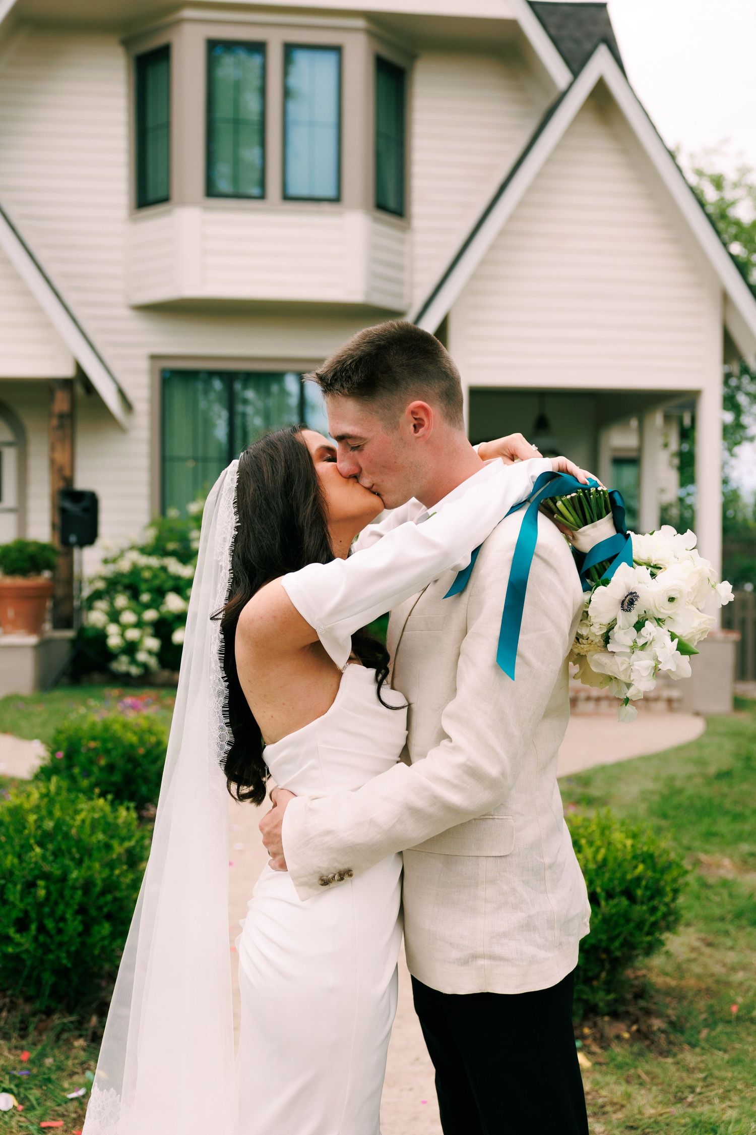 Romantic wedding couple embraces in front of a white house with green trim and beautiful landscaping.