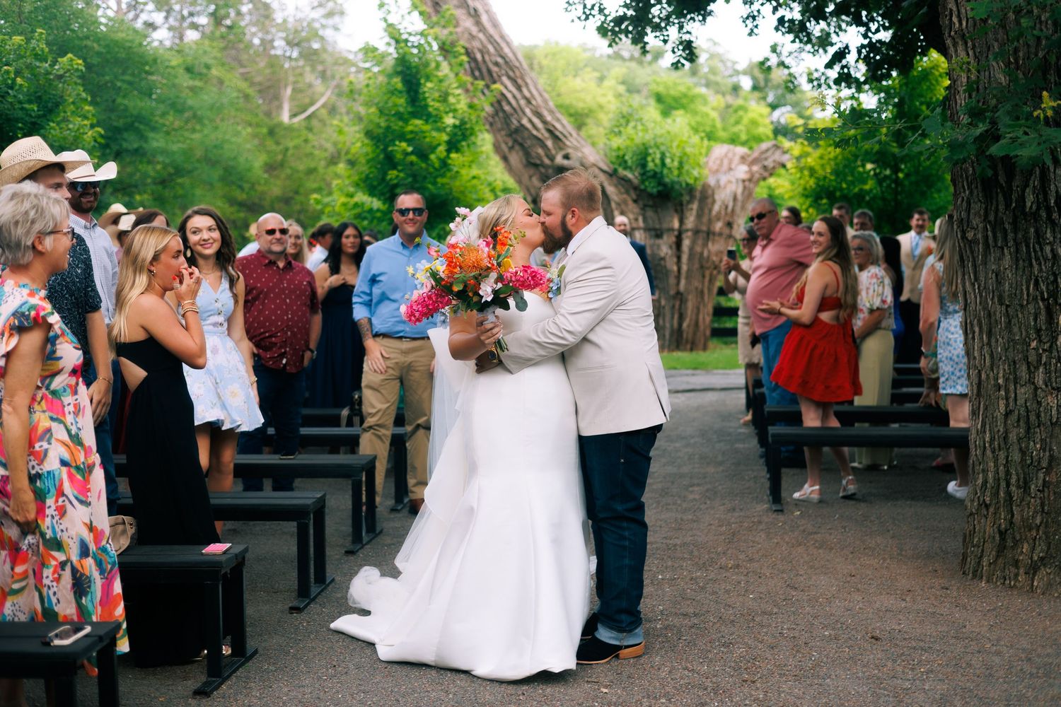 Outdoor wedding celebration with guests seated on benches surrounded by trees.