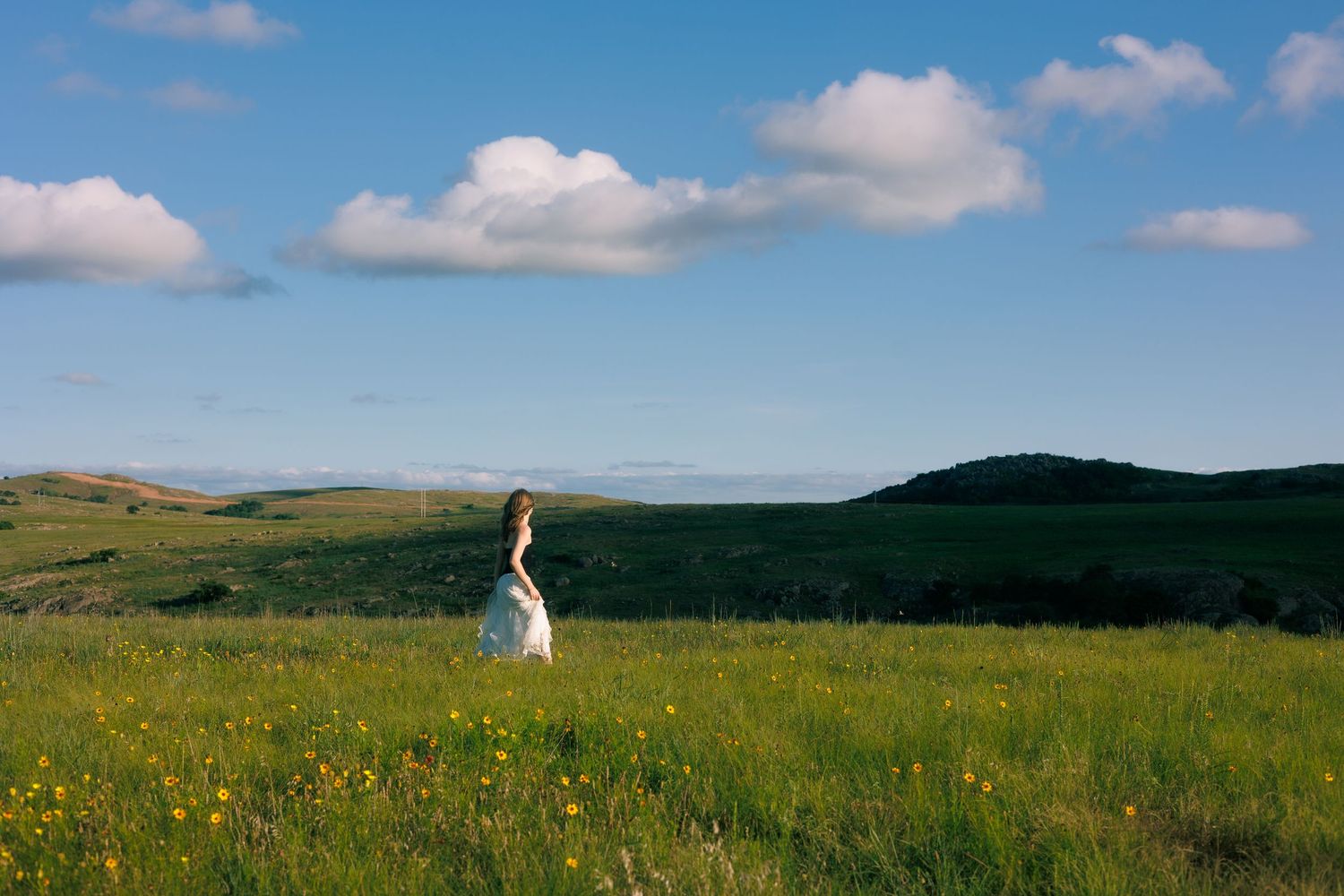 A figure in white stands alone in a vast meadow of wildflowers under a blue summer sky.