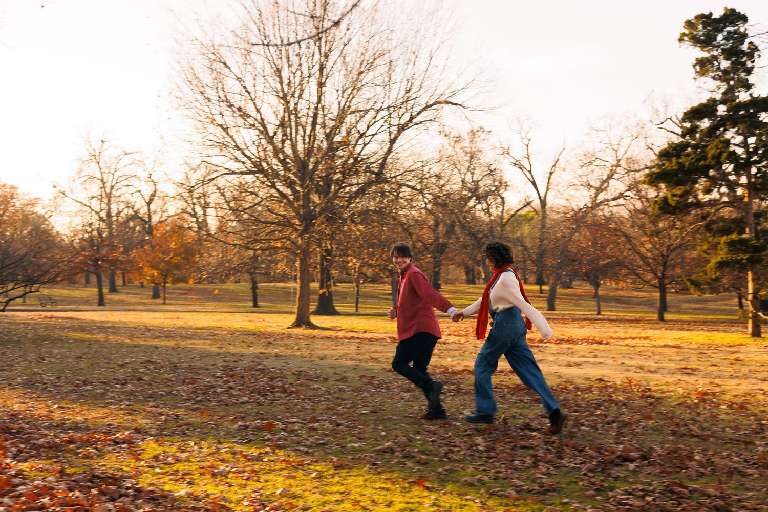 People walking through a park filled with fallen autumn leaves at sunset.