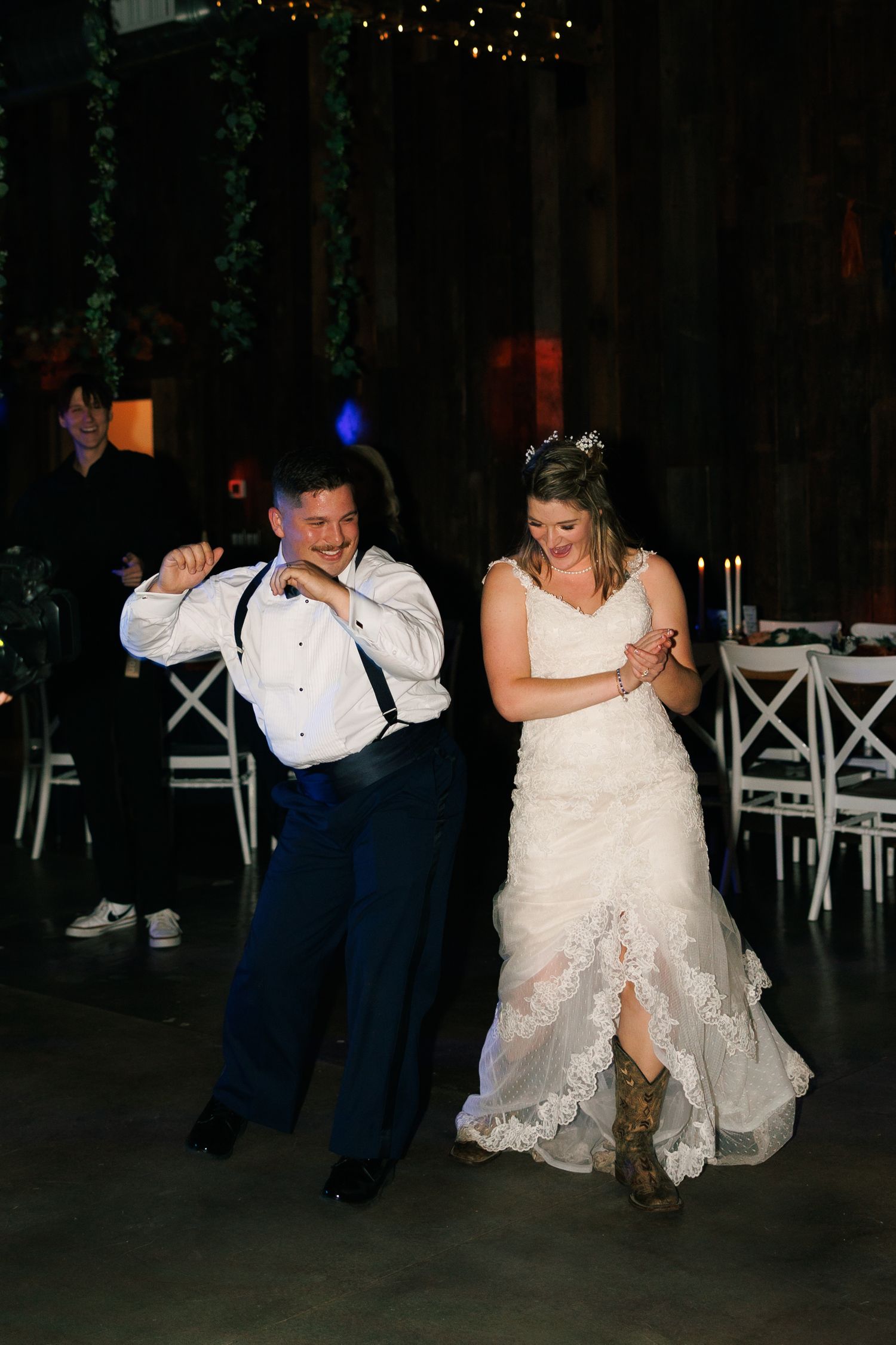 Wedding couple dances energetically at indoor reception with white chairs visible.