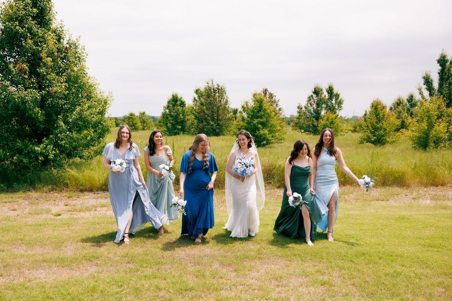 Wedding party in blue and white dresses sitting casually on grass in a sunny outdoor setting.