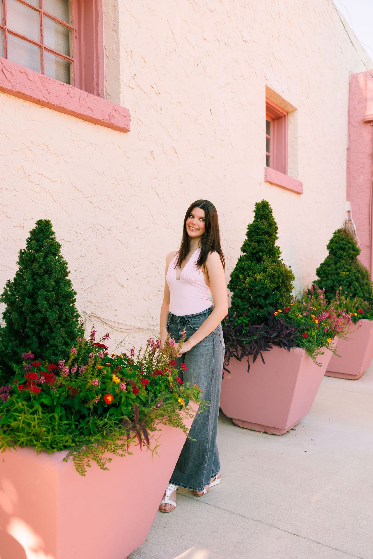 Person in casual outfit stands by pink stucco wall with potted evergreens and flowers.