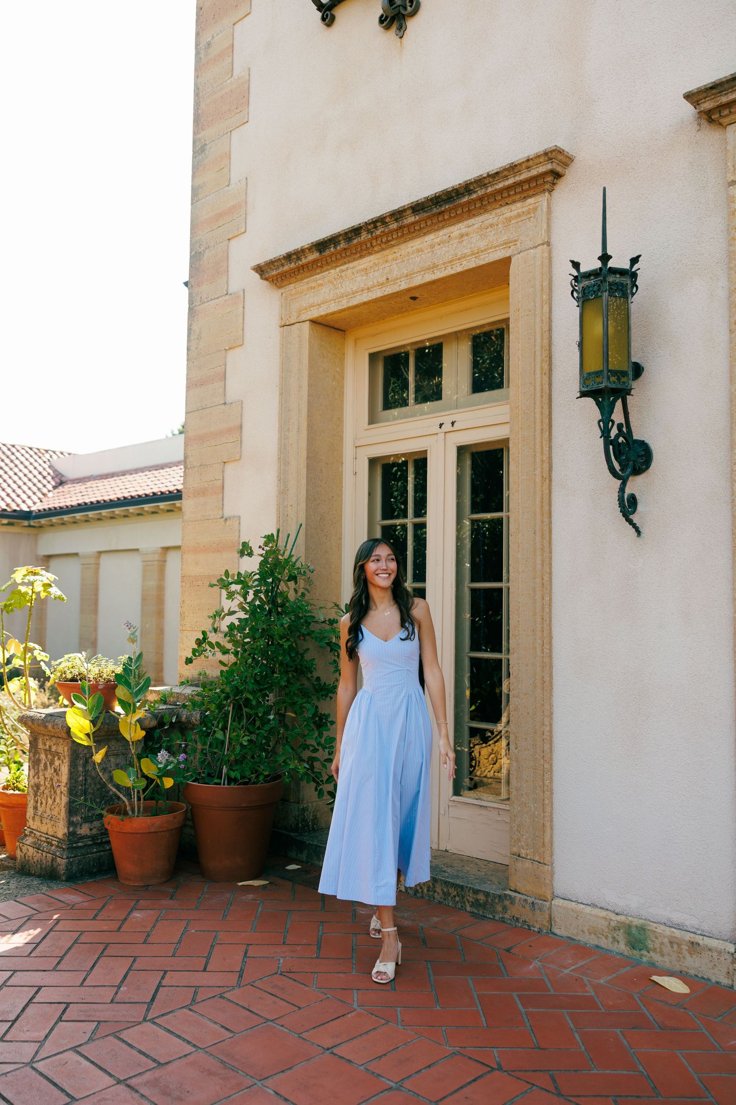 Woman in blue dress poses on terracotta patio near Spanish-style building.