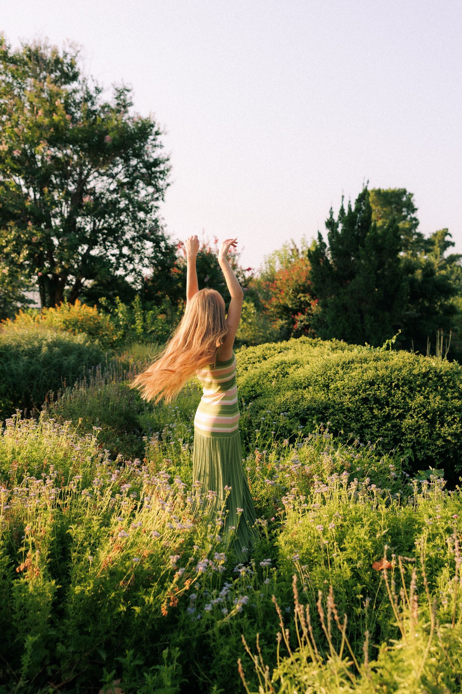 Woman in green striped top and skirt dancing with arms raised in a lush garden surrounded by blooming flowers and trees.