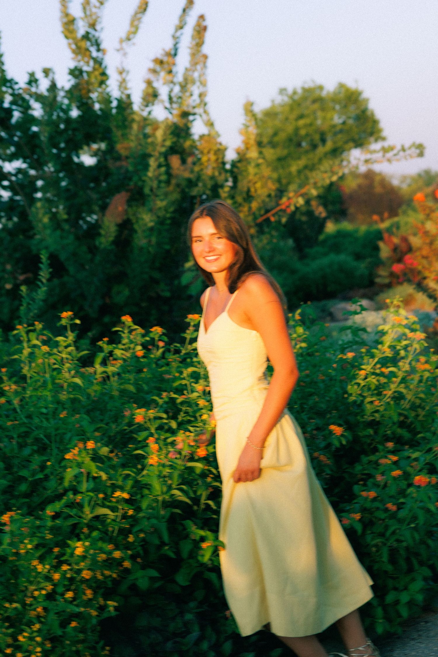 Young woman in yellow sundress smiling among colorful garden flowers at golden hour sunset.