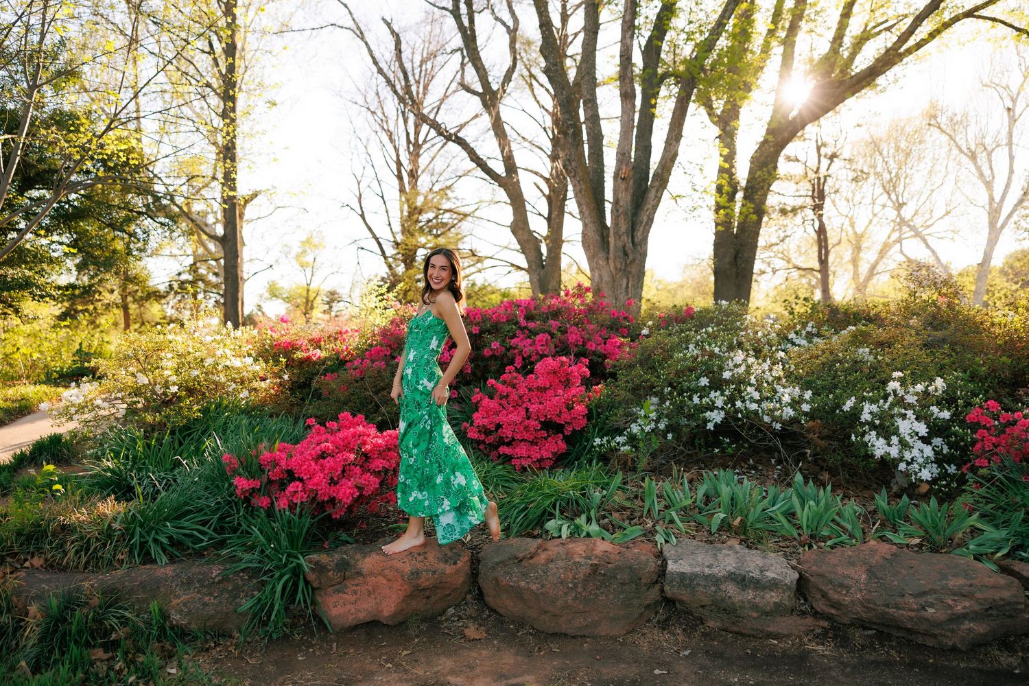 Woman in green floral dress posing among vibrant azalea blooms in a sunlit garden with trees in background.