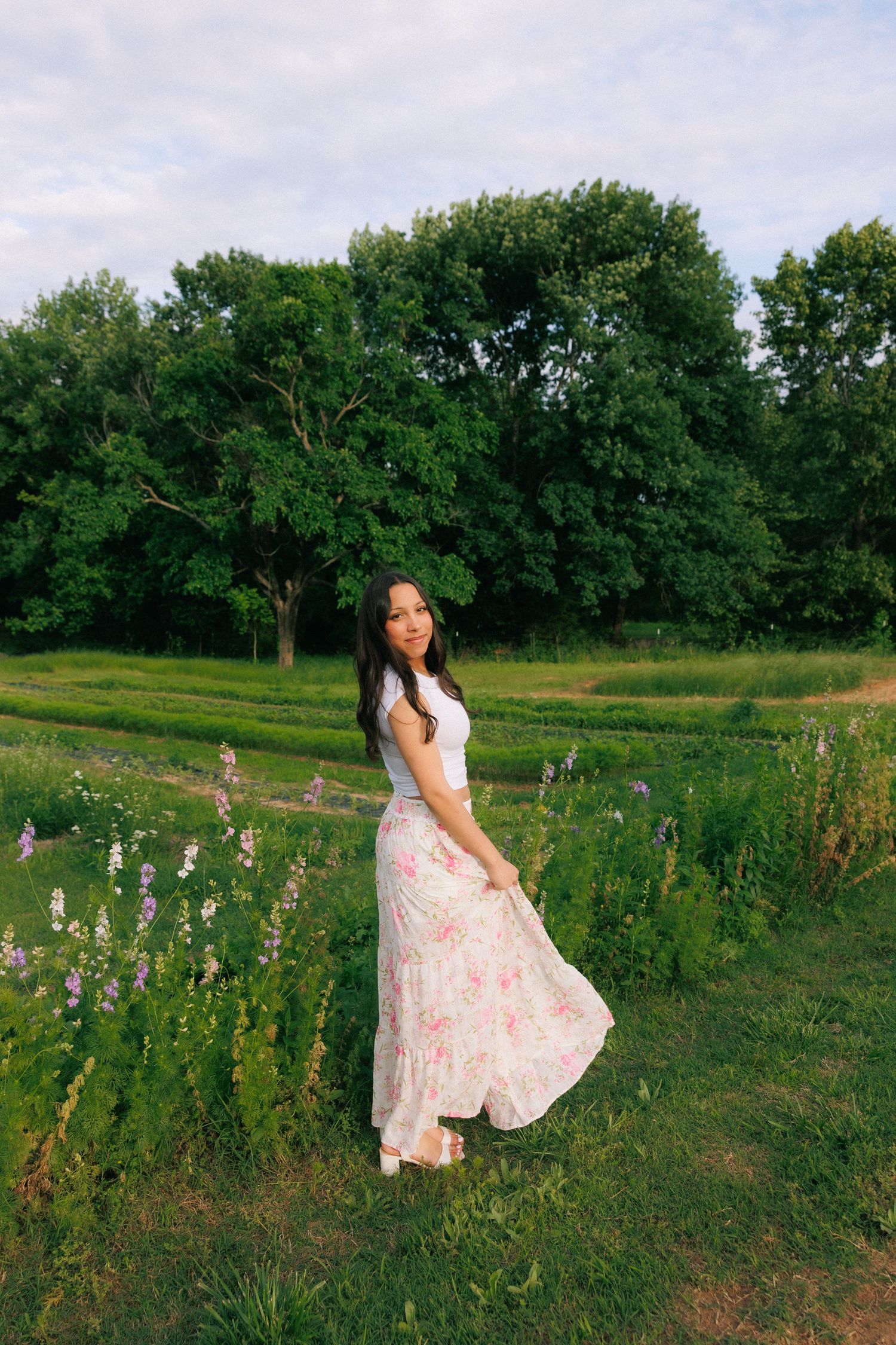 Young woman in floral maxi skirt and white top posing in a lush green field with wildflowers and trees.