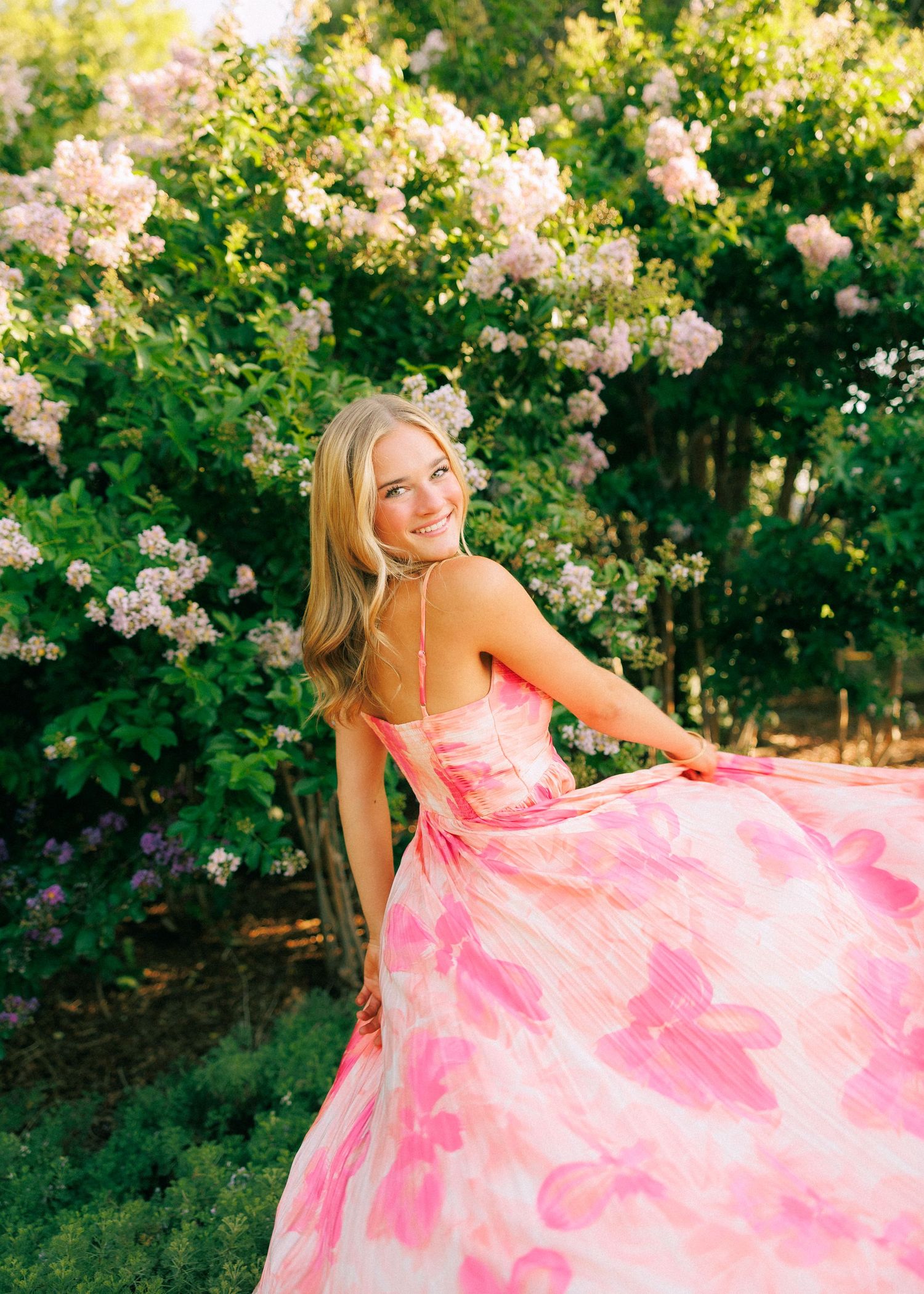 Smiling blonde woman twirling in a pink floral gown outdoors surrounded by lush green bushes with soft pink blooms.