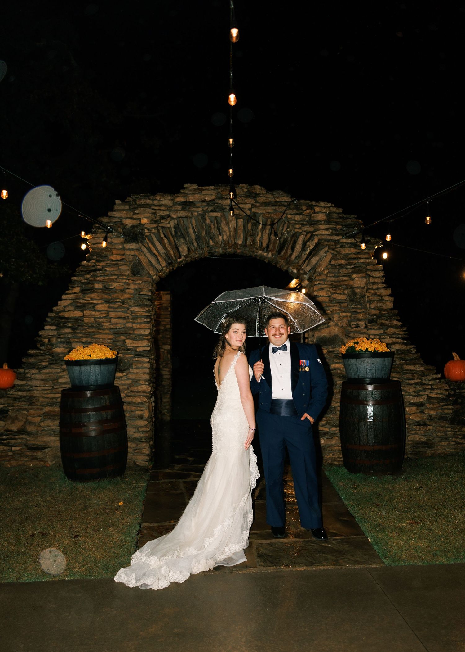 Couple poses for nighttime photos in front of stone archway with barrels.