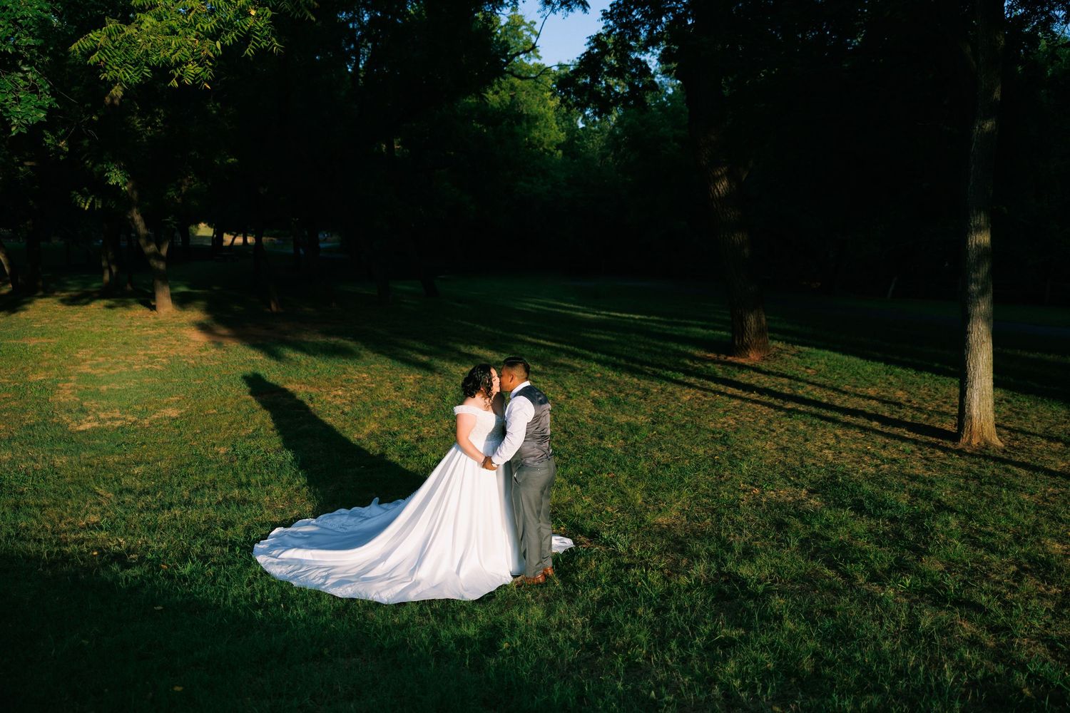 A wedding couple shares romantic moments on a sunlit grassy hillside.