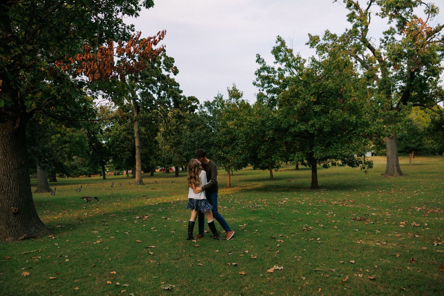 Two people walking together through a park with scattered autumn leaves on the ground.